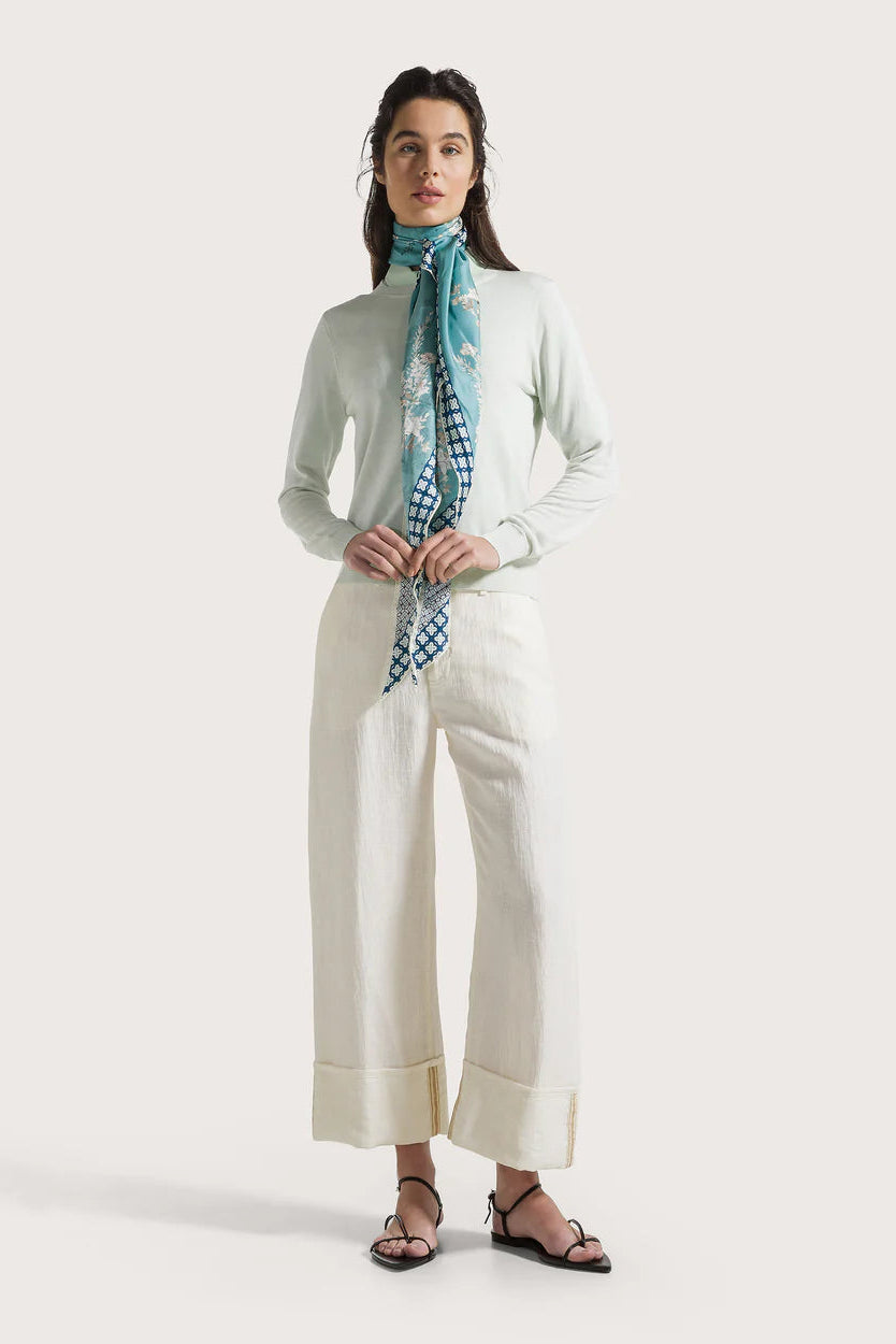 Woman wearing a light blue sweater, white pants, and a patterned scarf on a white background.