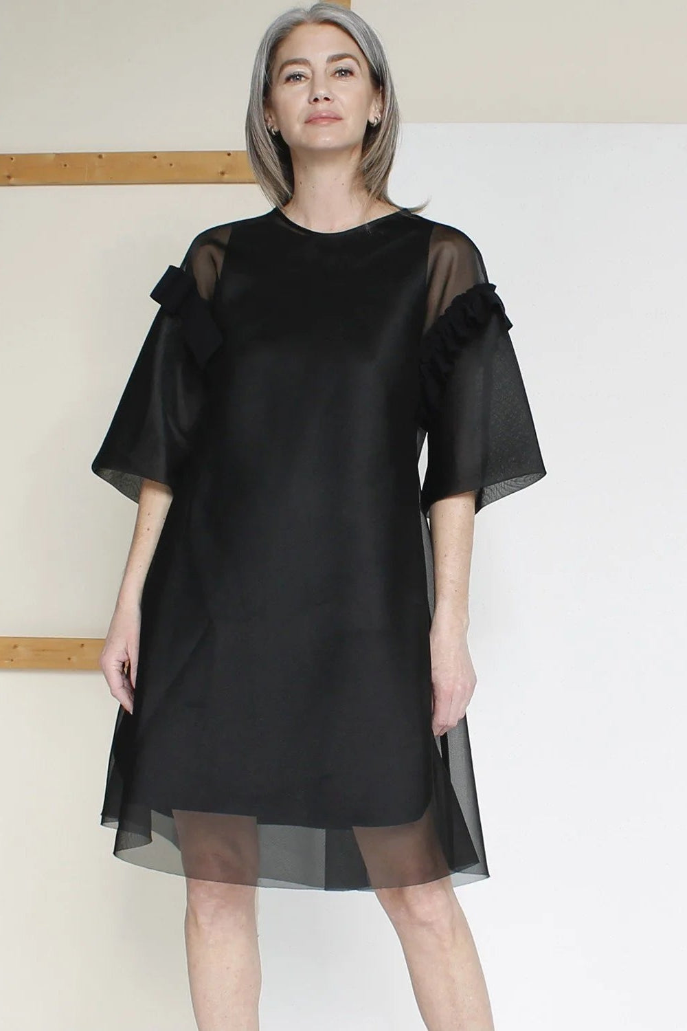 Woman wearing a black dress with sheer sleeves against a neutral background.