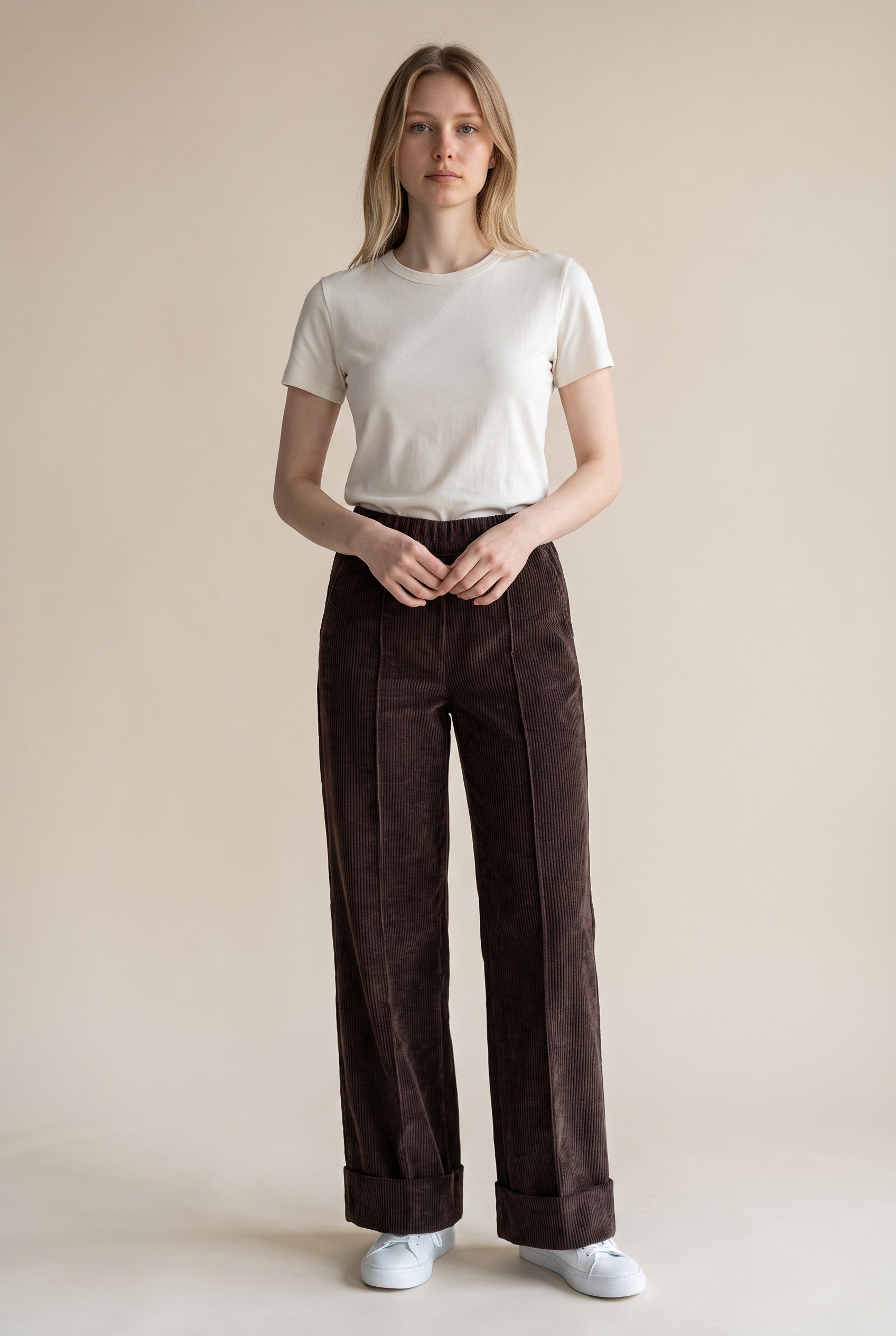 A woman stands against a plain beige studio background wearing a white short-sleeve crewneck t-shirt, dark brown wide-leg corduroy pants, and white sneakers.