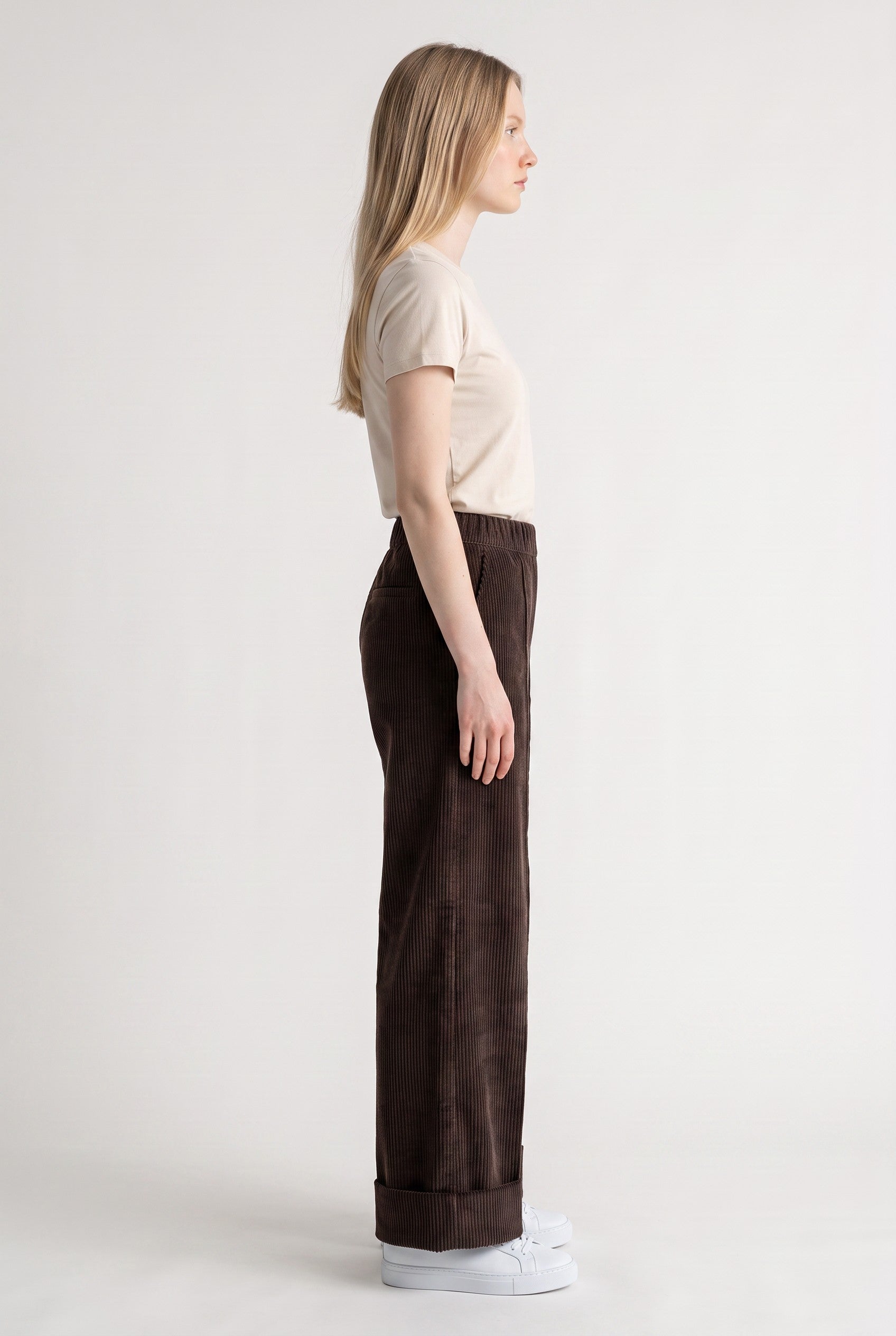 A woman shown in profile wearing dark brown wide-leg corduroy pants, a beige short-sleeve t-shirt, and white sneakers against a plain light gray studio background.