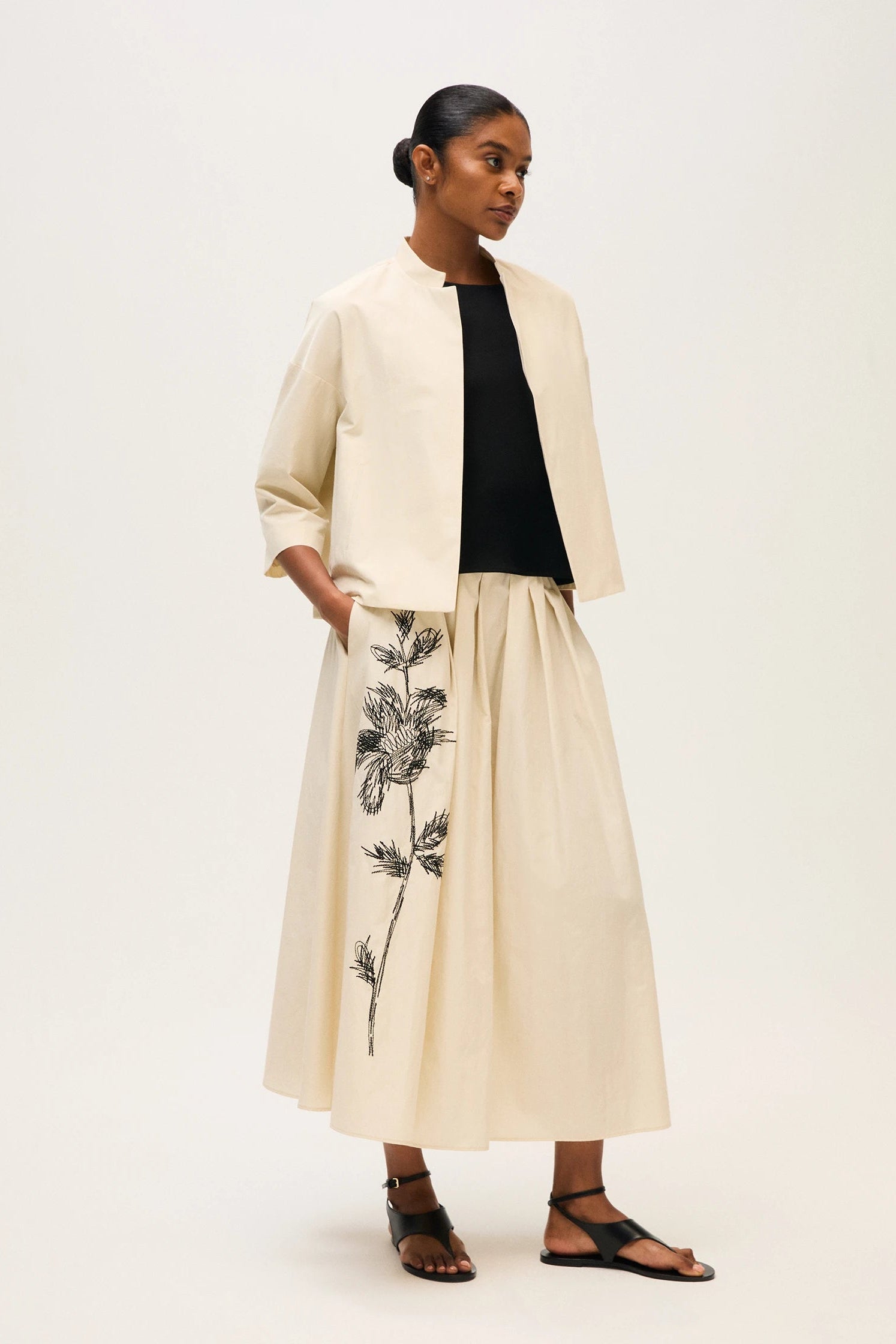 Woman wearing a beige coat over a black top and beige skirt with floral embroidery on a white background.