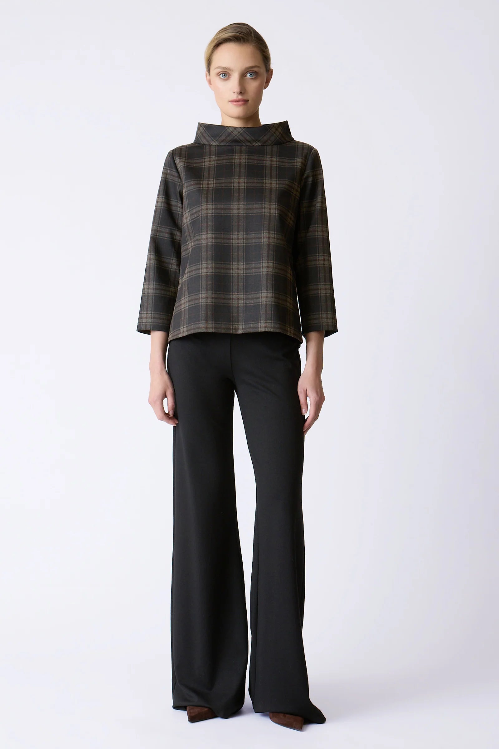 A model stands against a light gray studio background wearing a dark brown and black plaid cropped long-sleeve top with a high mock neck, paired with black wide-leg trousers.