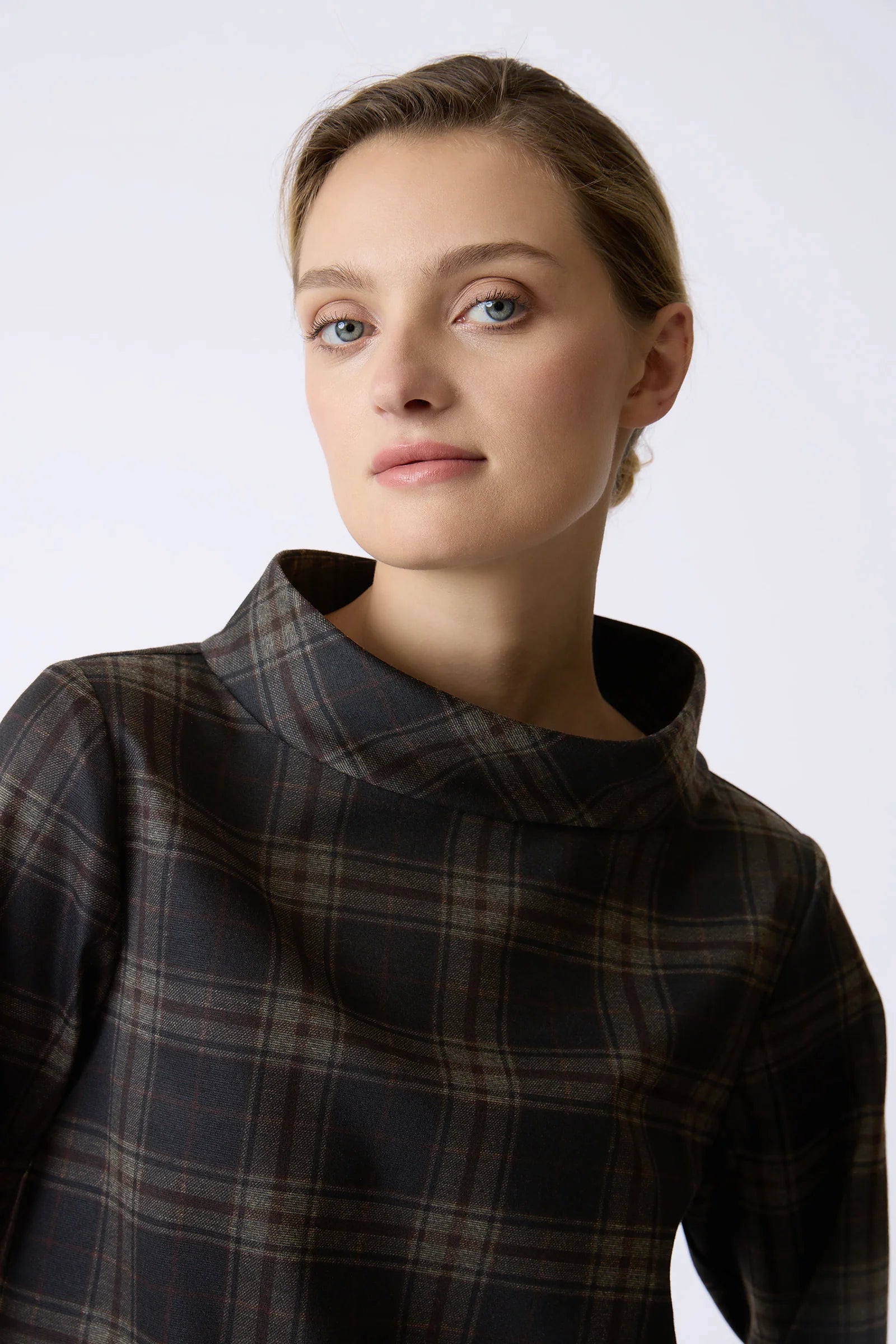 A close-up portrait of a woman wearing a dark plaid high-neck top against a plain light gray studio background.