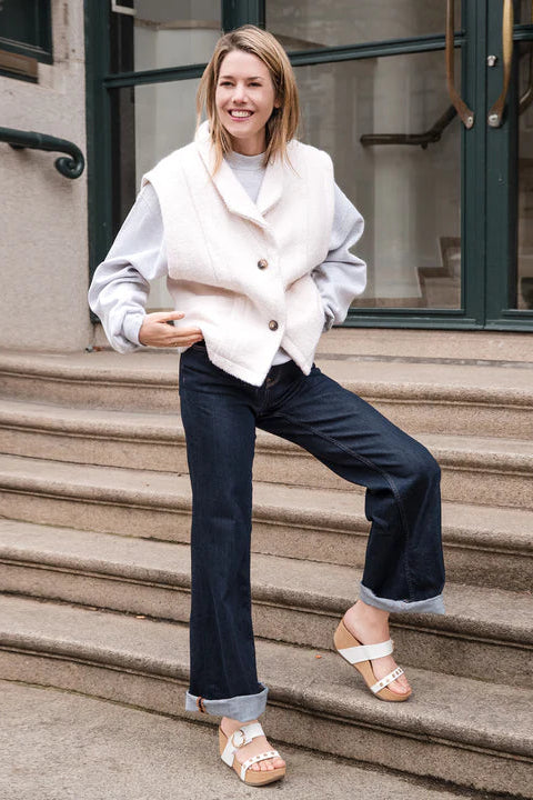 Woman in a white vest and blue jeans and white platfrom sndals standing on steps outside a building.