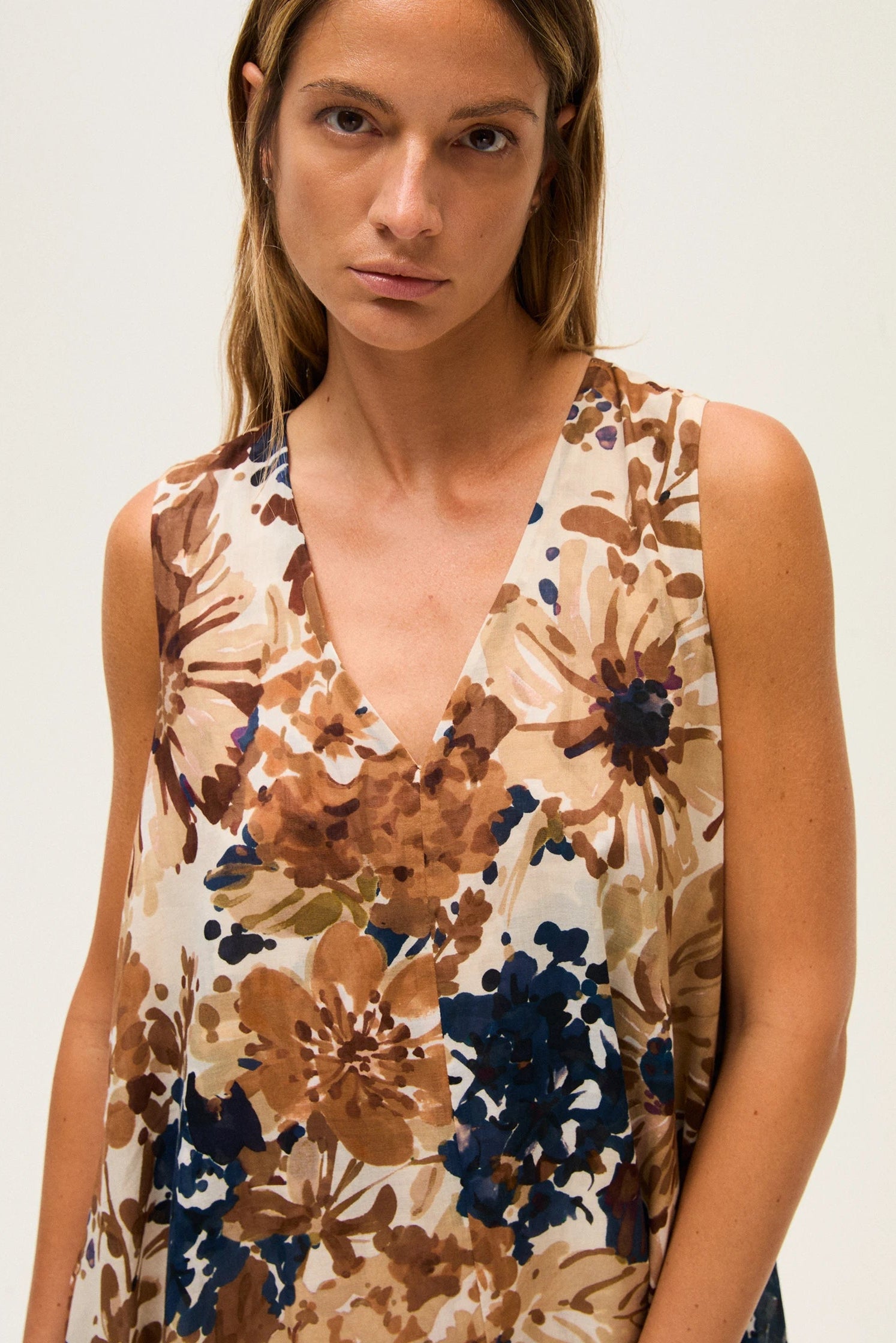 Close-up of a woman wearing a sleeveless floral dress with brown, blue, and beige colors on a white background.