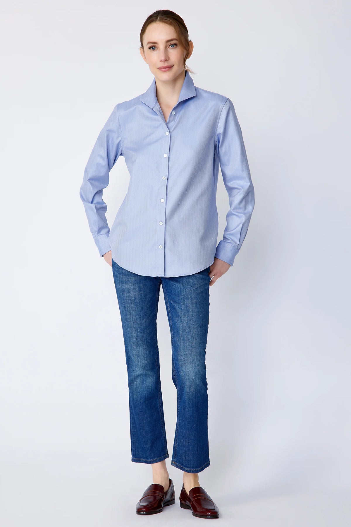 A woman modeling a light blue long-sleeve button-down shirt with blue jeans and brown leather loafers, standing against a light gray studio background.