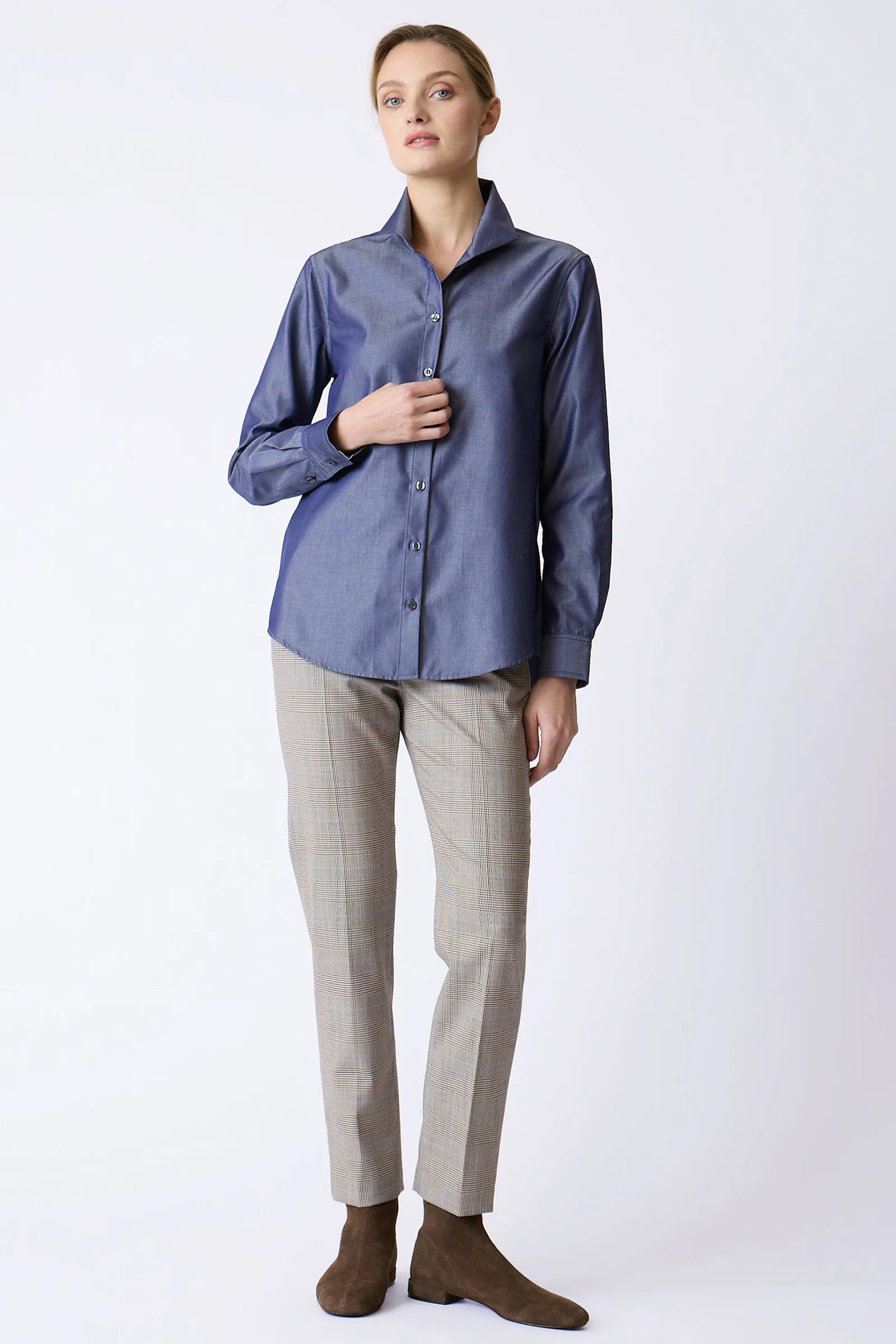 A woman modeling a slate blue long-sleeve button-up shirt paired with beige trousers and brown suede boots, standing against a light gray studio background.