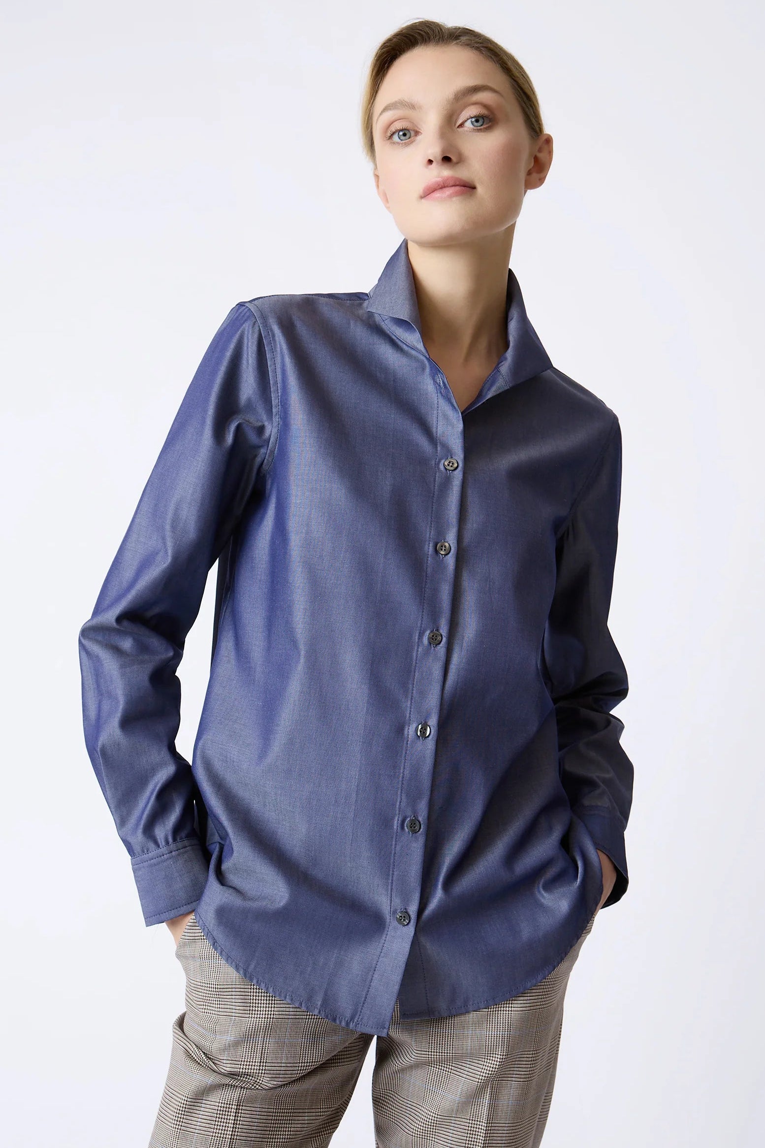 A woman wearing a long-sleeve button-up shirt in a dusty slate blue color, paired with beige trousers, photographed against a light gray studio background.