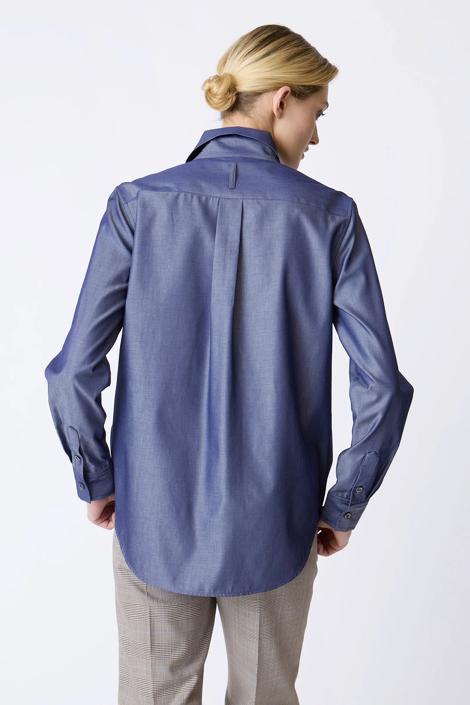 A woman shown from the back wearing a slate blue long-sleeve button-down shirt and light beige trousers, photographed against a clean white studio background.
