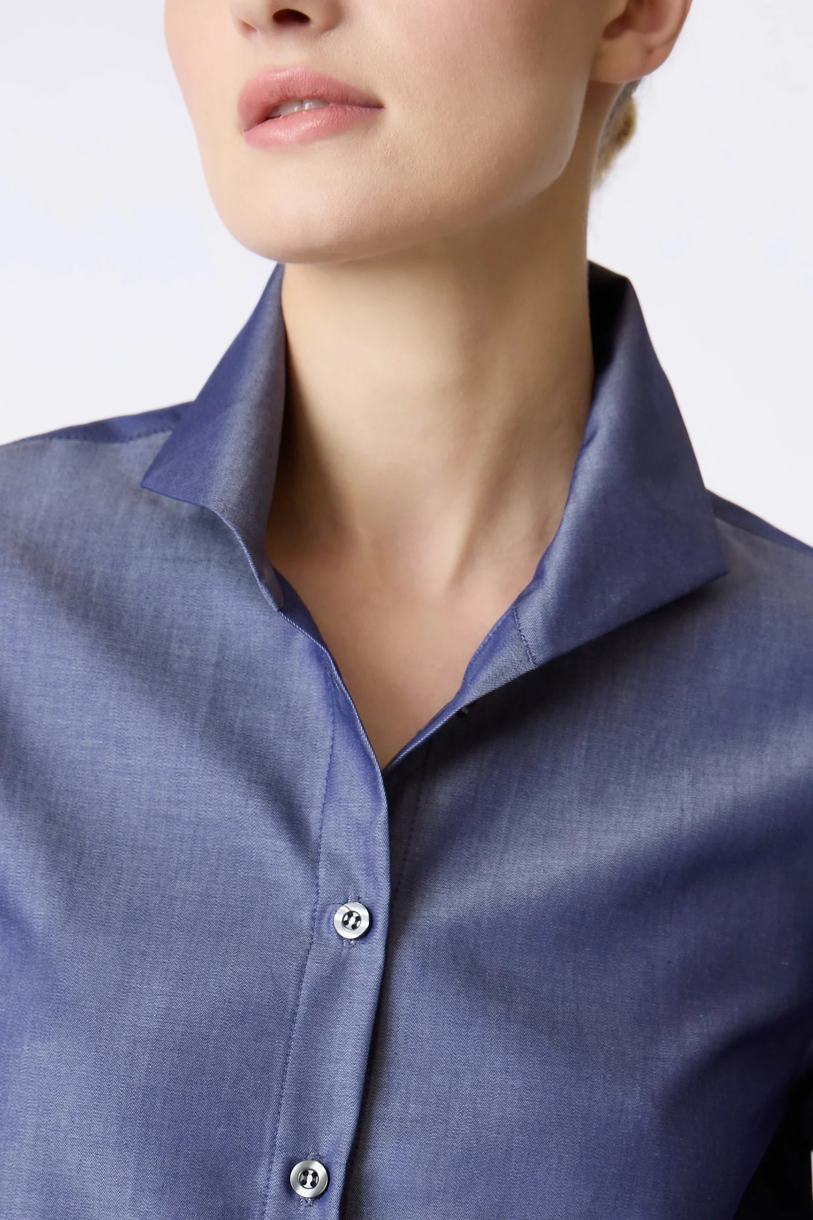 Close-up of a woman wearing a blue collared button-up shirt, showing the collar, chest area, and part of the face against a light studio background.