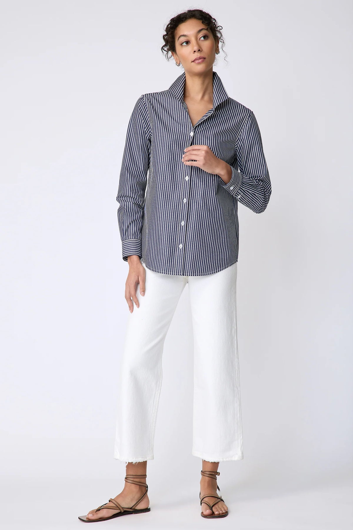A woman modeling a navy and white vertically striped button-down shirt with white cropped pants and brown strappy sandals, standing against a light gray studio background.