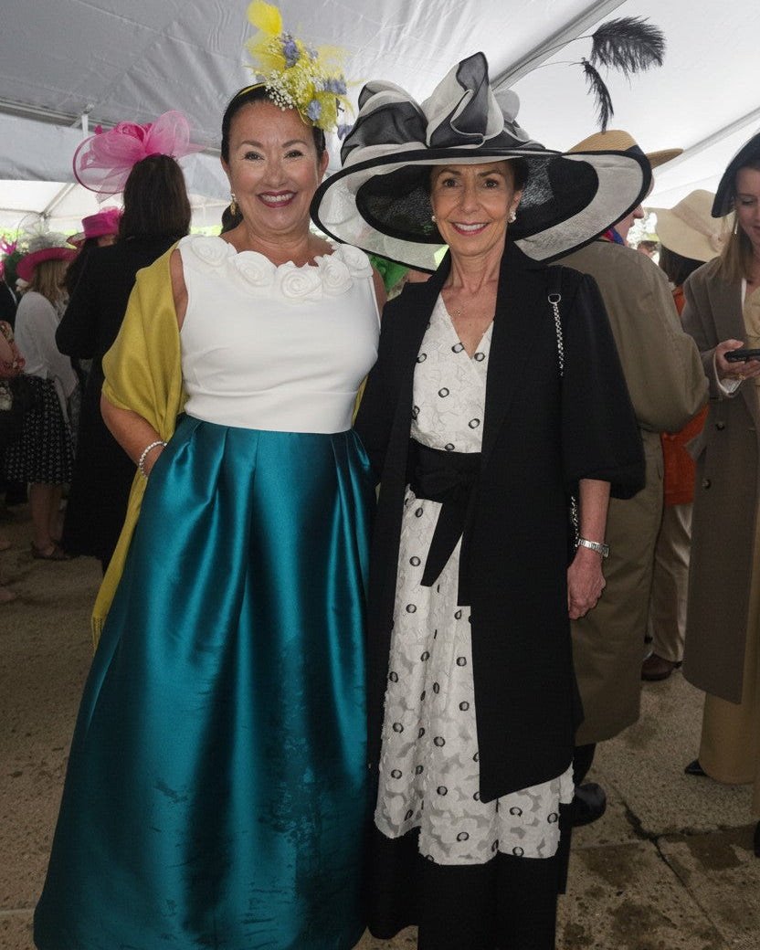 Two women in elaborate hats and dresses standing together at an event with a crowd in the background.