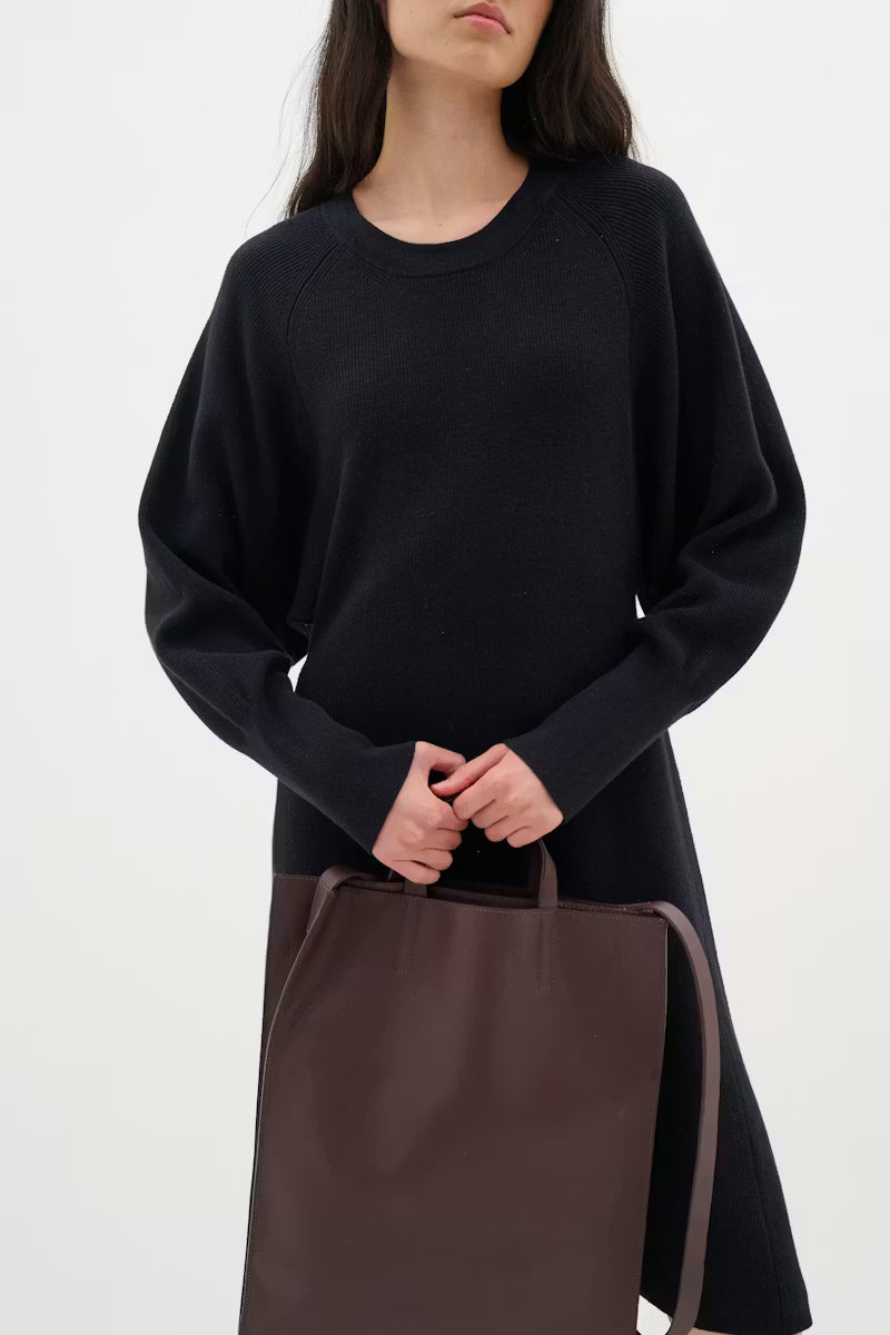 Woman wearing a black sweater dress with a brown bag on a white background.