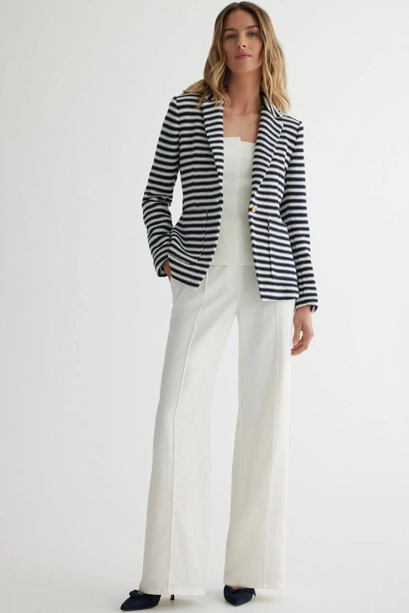Woman wearing a navy and white striped blazer over a white top and pants on a white background.