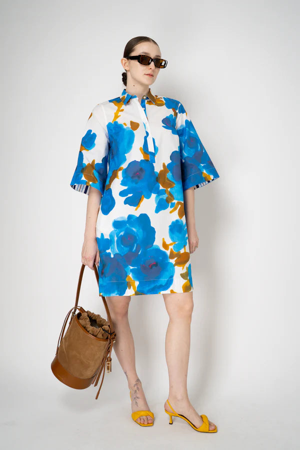 Woman wearing a floral dress with blue flowers, holding a brown bag on a white background.