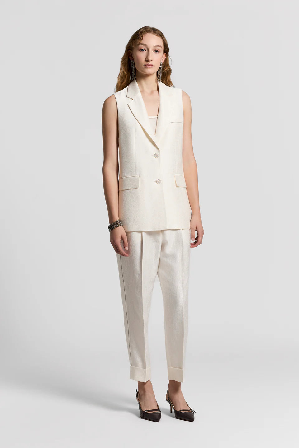 Woman wearing a white sleeveless blazer and white pants on a plain background.