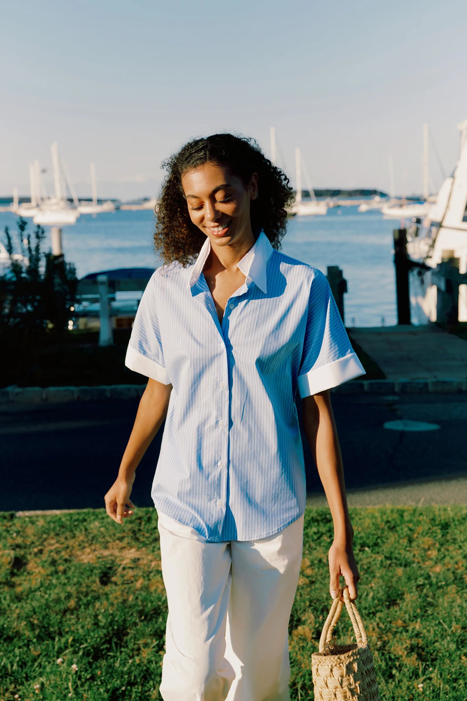 Woman in a light blue and white outfit standing on grass with boats in the background.