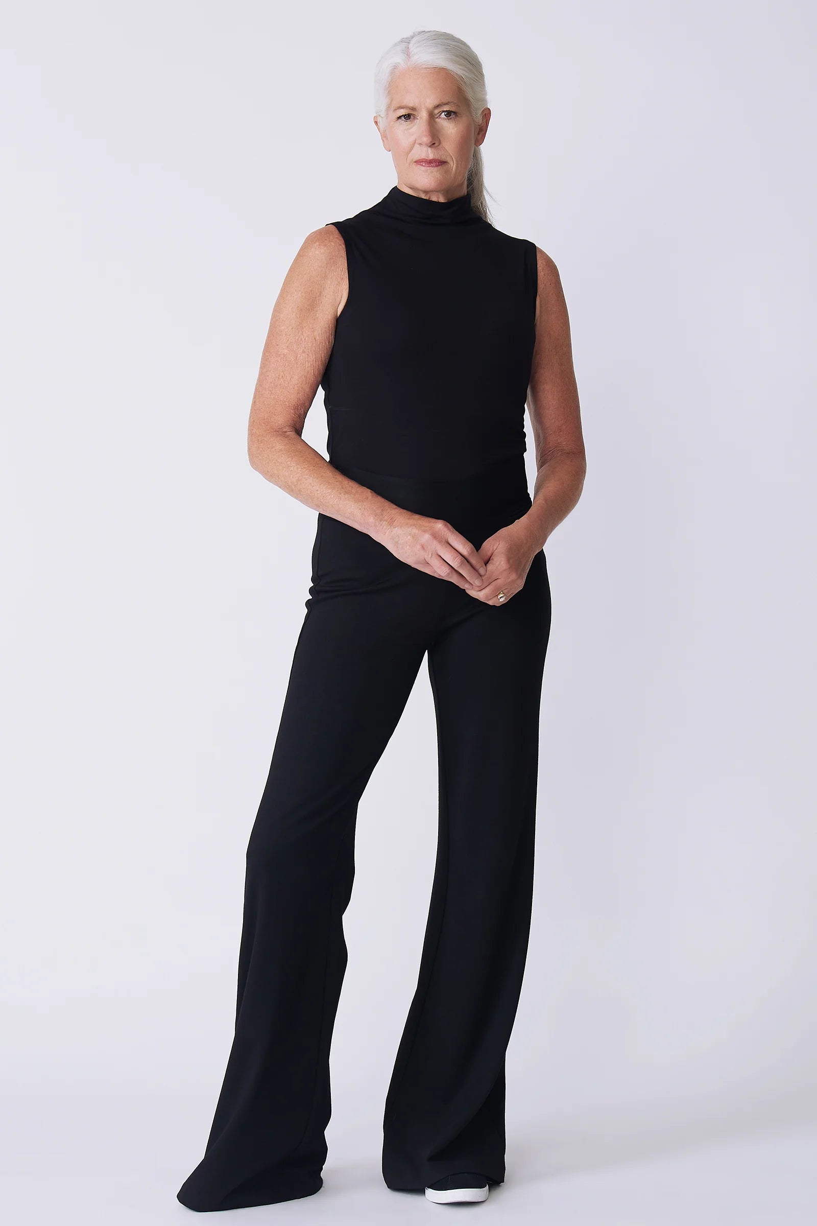 A woman with short gray hair models a black sleeveless mock-neck top and matching black wide-leg pants against a light gray studio background.