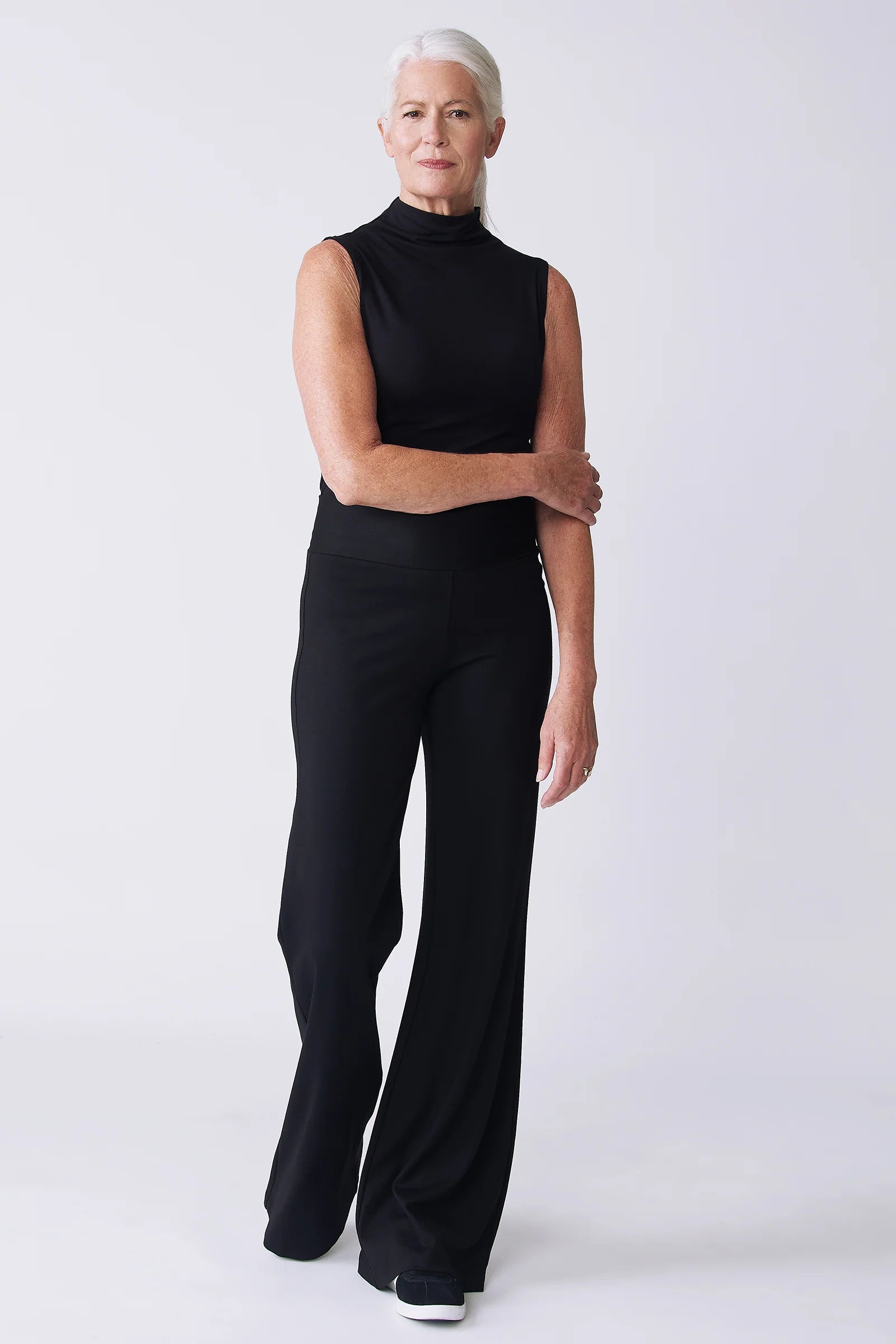 A woman with short white hair models a black sleeveless mock-neck top and matching black wide-leg pants against a light gray studio background.