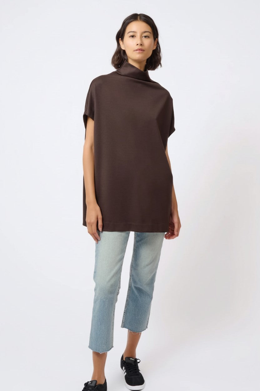 A woman modeling a dark brown short-sleeve tunic top with a high mock neck, paired with light-wash cropped jeans and black sneakers, against a plain white studio background.
