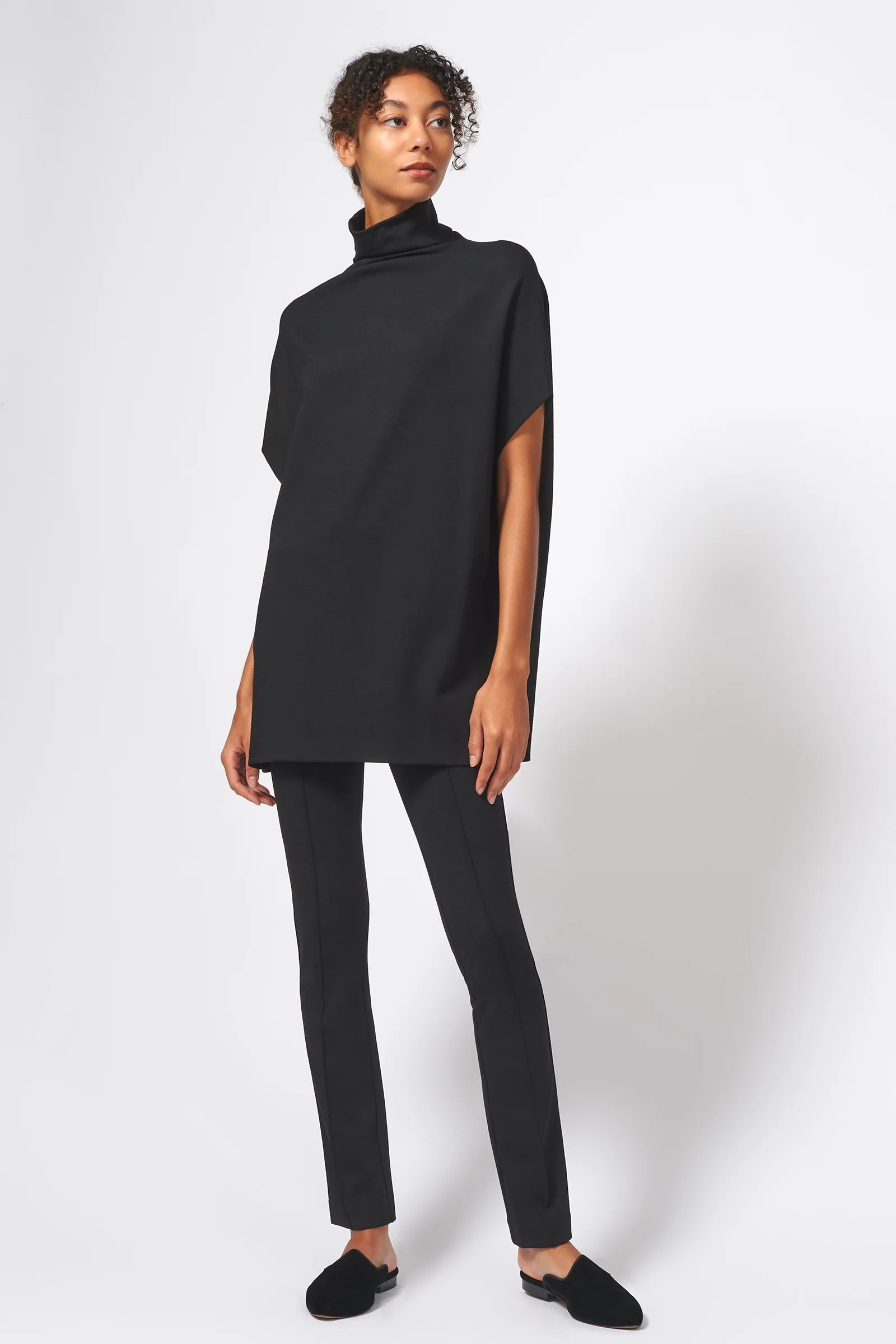 A model wearing a black oversized short-sleeve turtleneck top, black slim-fit pants, and black slip-on shoes, standing against a light gray studio background.