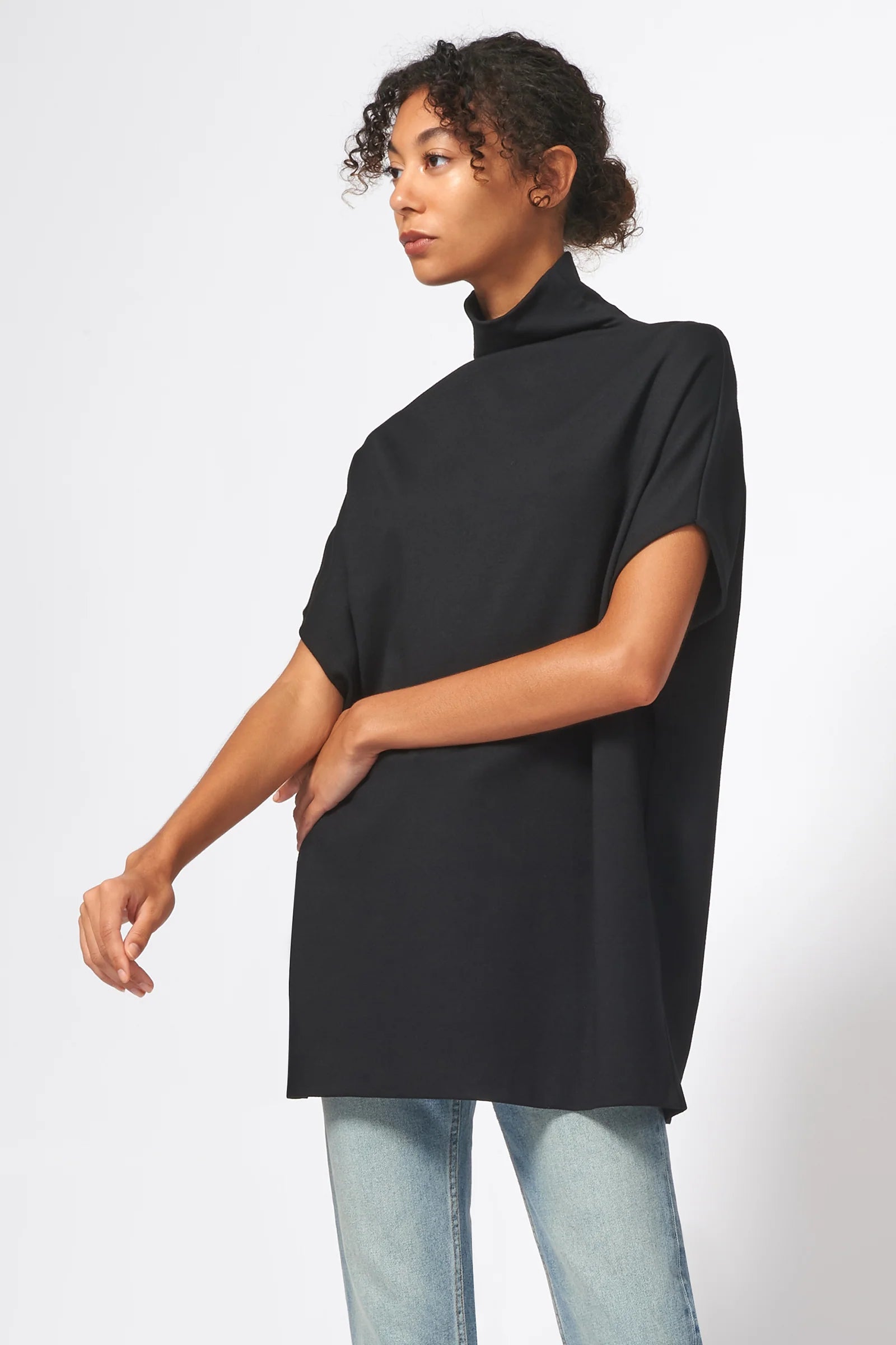 A woman modeling a black short-sleeve mock neck tunic top with light wash blue jeans, photographed against a clean white studio background.