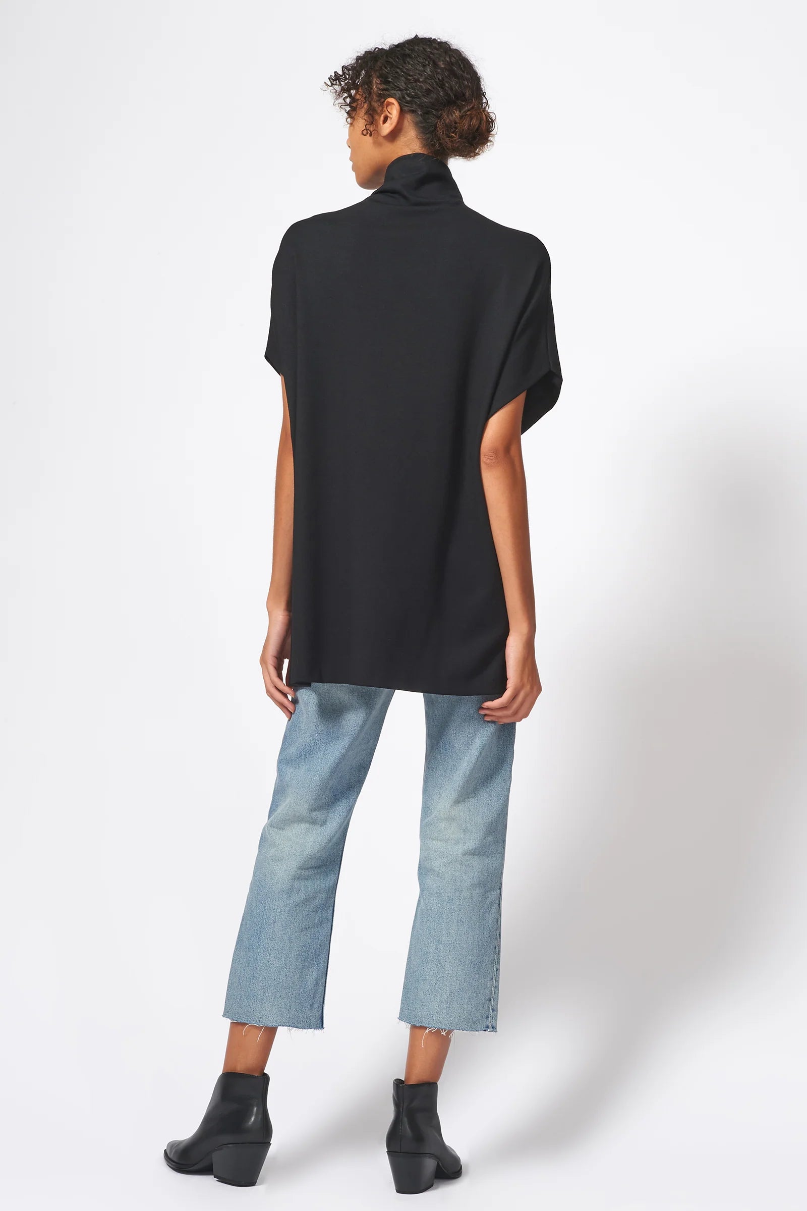 A model shown from the back wearing a black short-sleeve mock neck top, light-wash blue cropped jeans, and black ankle boots against a white studio background.