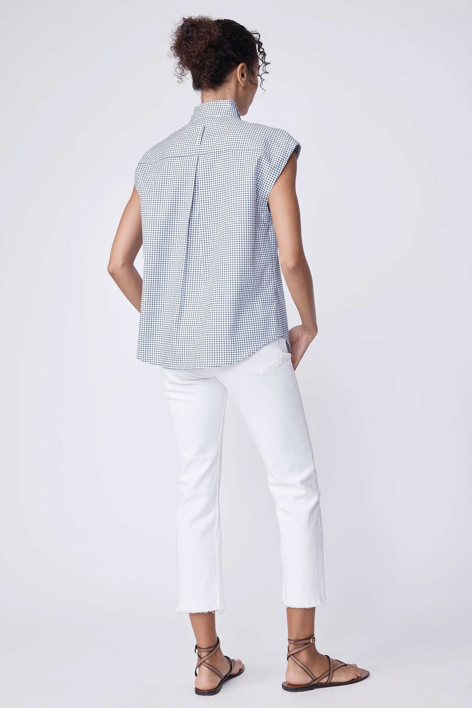 Back of a woman wearing a blue checkered sleeveless shirt and white pants on a white background.