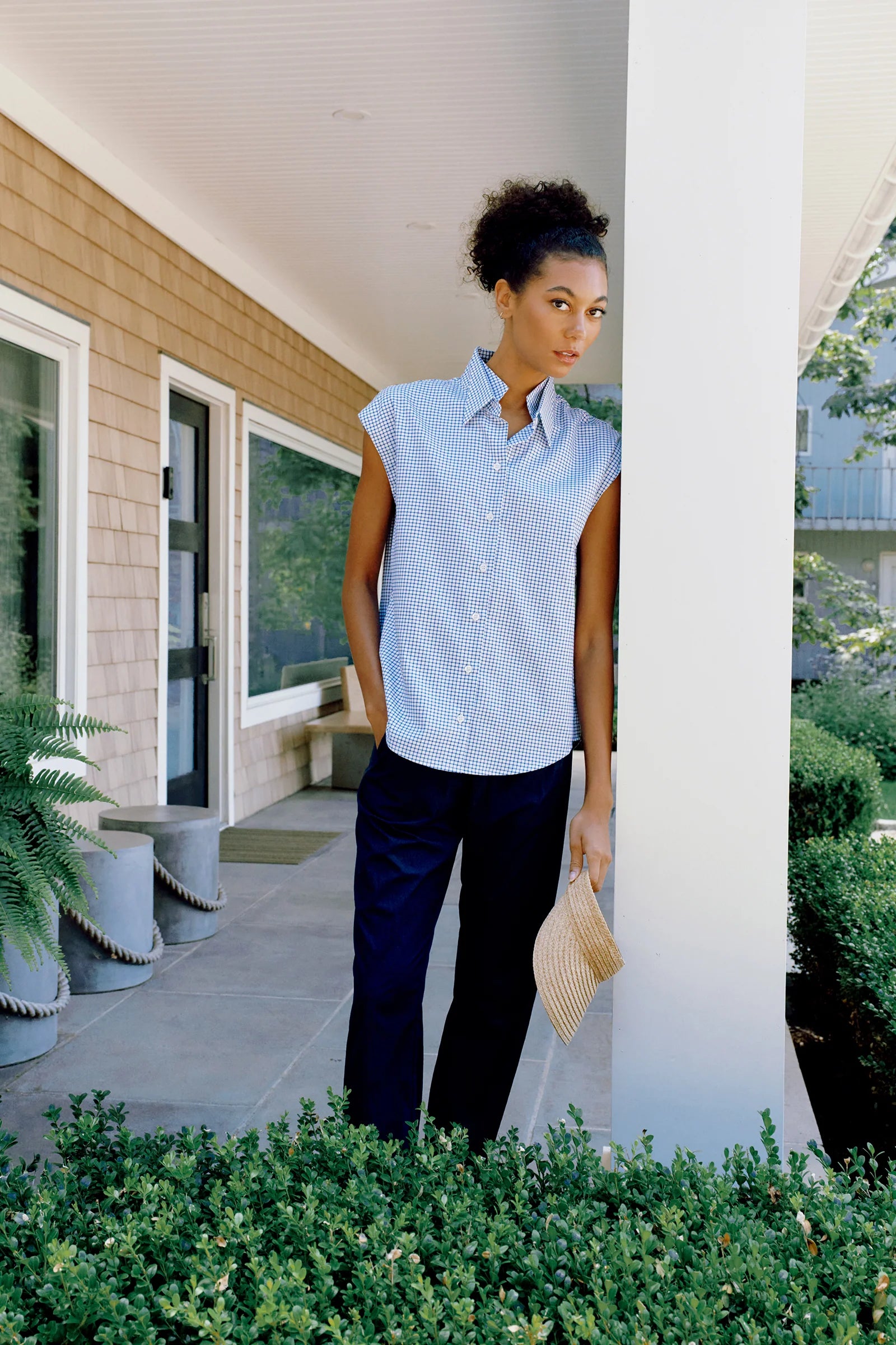 Woman standing on a porch wearing a light blue sleeveless shirt and dark pants.