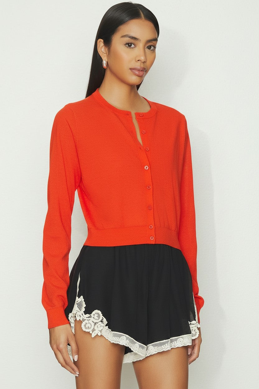 Woman wearing a orange cardigan over a black outfit with lace details on a white background.
