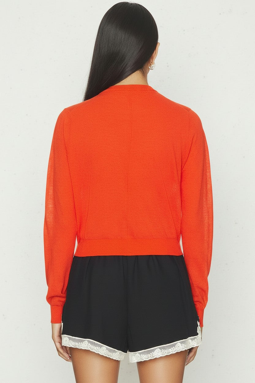 The back of a woman wearing a orange cardigan and black shorts on a white background.