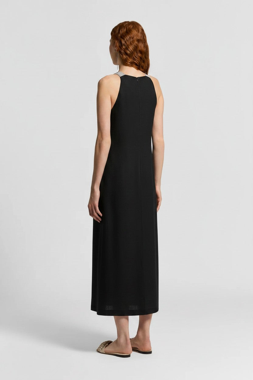 The back of a woman wearing a black linen dress and gold sandals on a plain background.