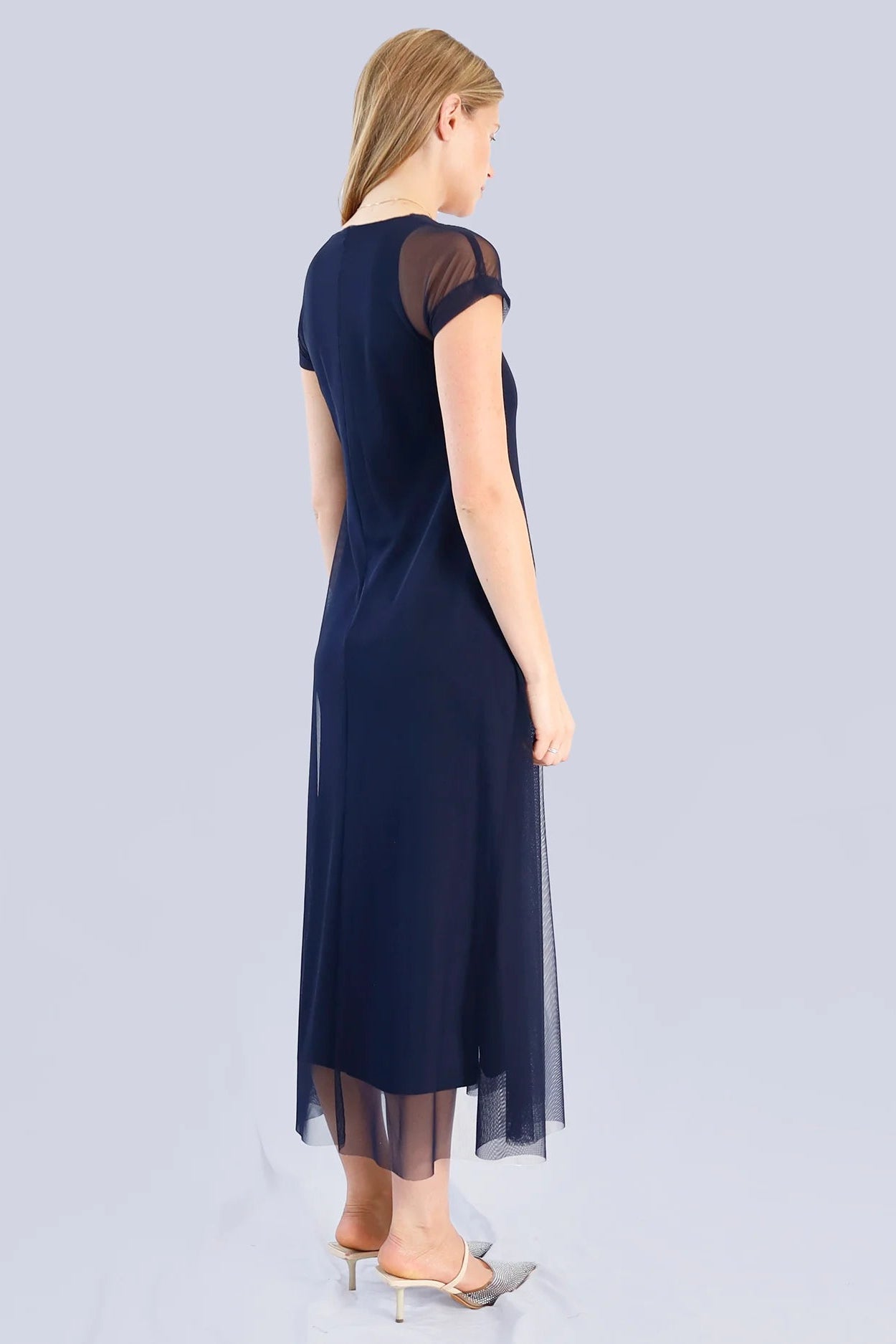 The back of a woman wearing a navy blue dress with sheer sleeves on a light gray background.