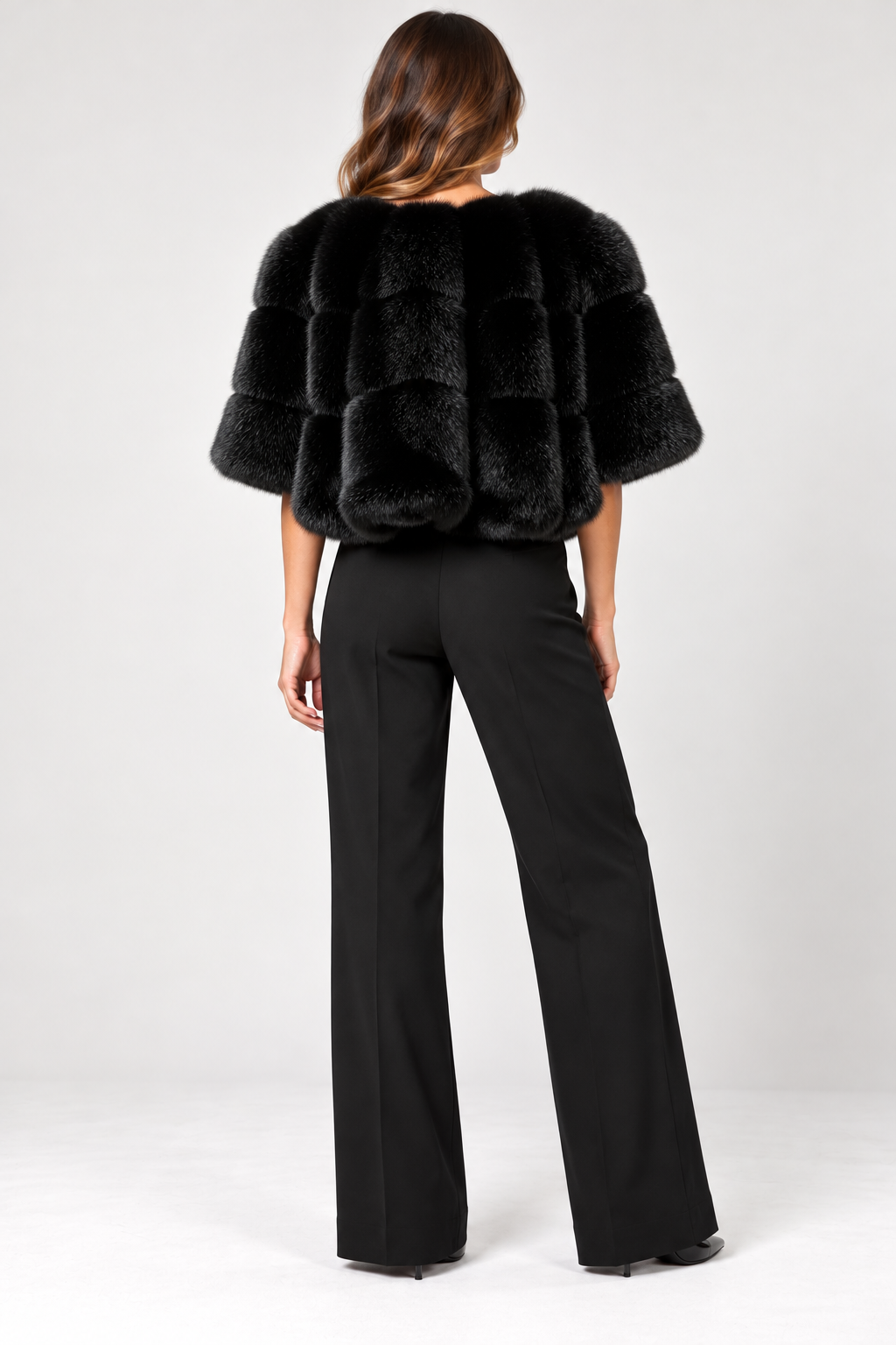 Back view of model wearing the Suprema Black Mink Fur Cropped Capelet, featuring luxurious genuine black mink fur, a rounded neckline, voluminous elbow-length sleeves, horizontal fur panels, and a cropped silhouette. Styled with tailored black wide-leg trousers and pointed-toe heels against a neutral studio background.