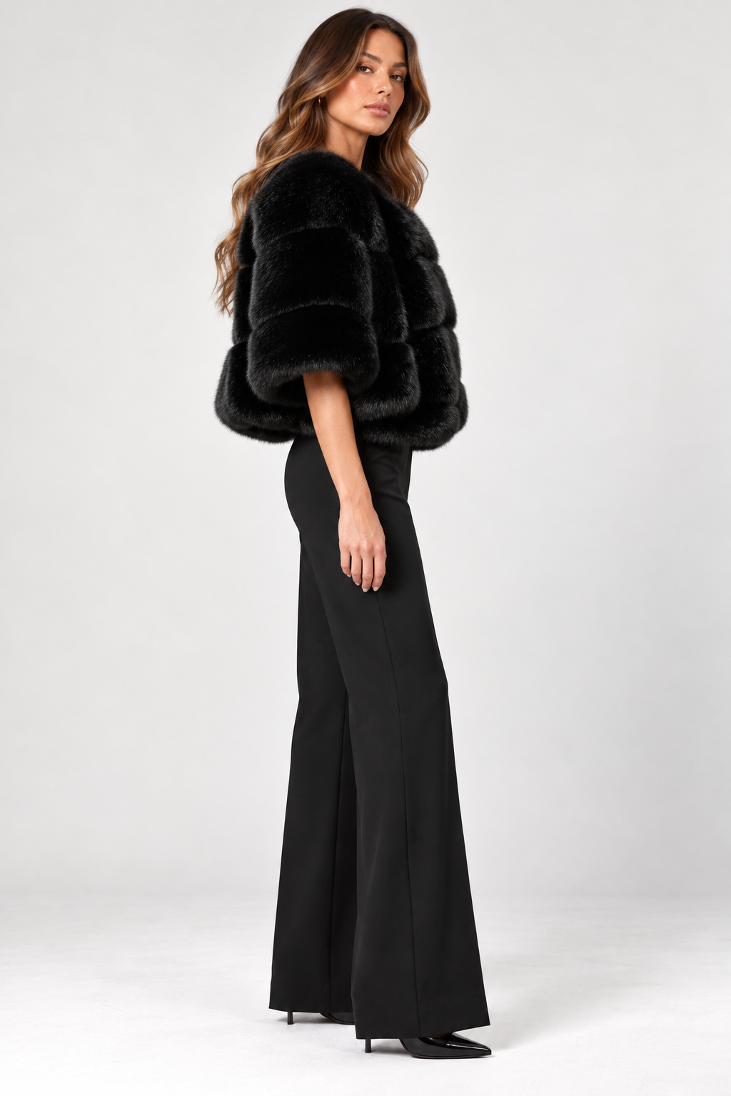 Side view of model wearing the Suprema Black Mink Fur Cropped Capelet, featuring luxurious genuine black mink fur, a rounded neckline, voluminous elbow-length sleeves, layered fur panels, and a cropped silhouette. Styled with tailored black wide-leg trousers and pointed-toe heels against a neutral studio background.