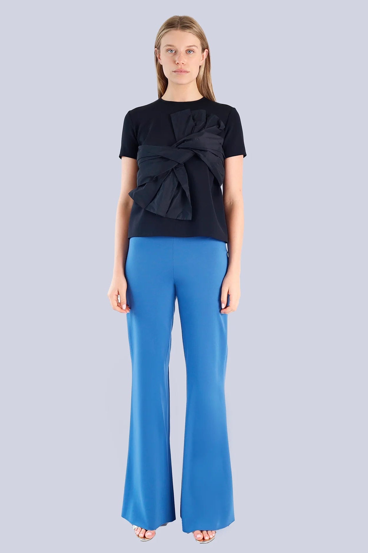 Woman wearing a black top with a bow detail and blue pants on a light gray background.