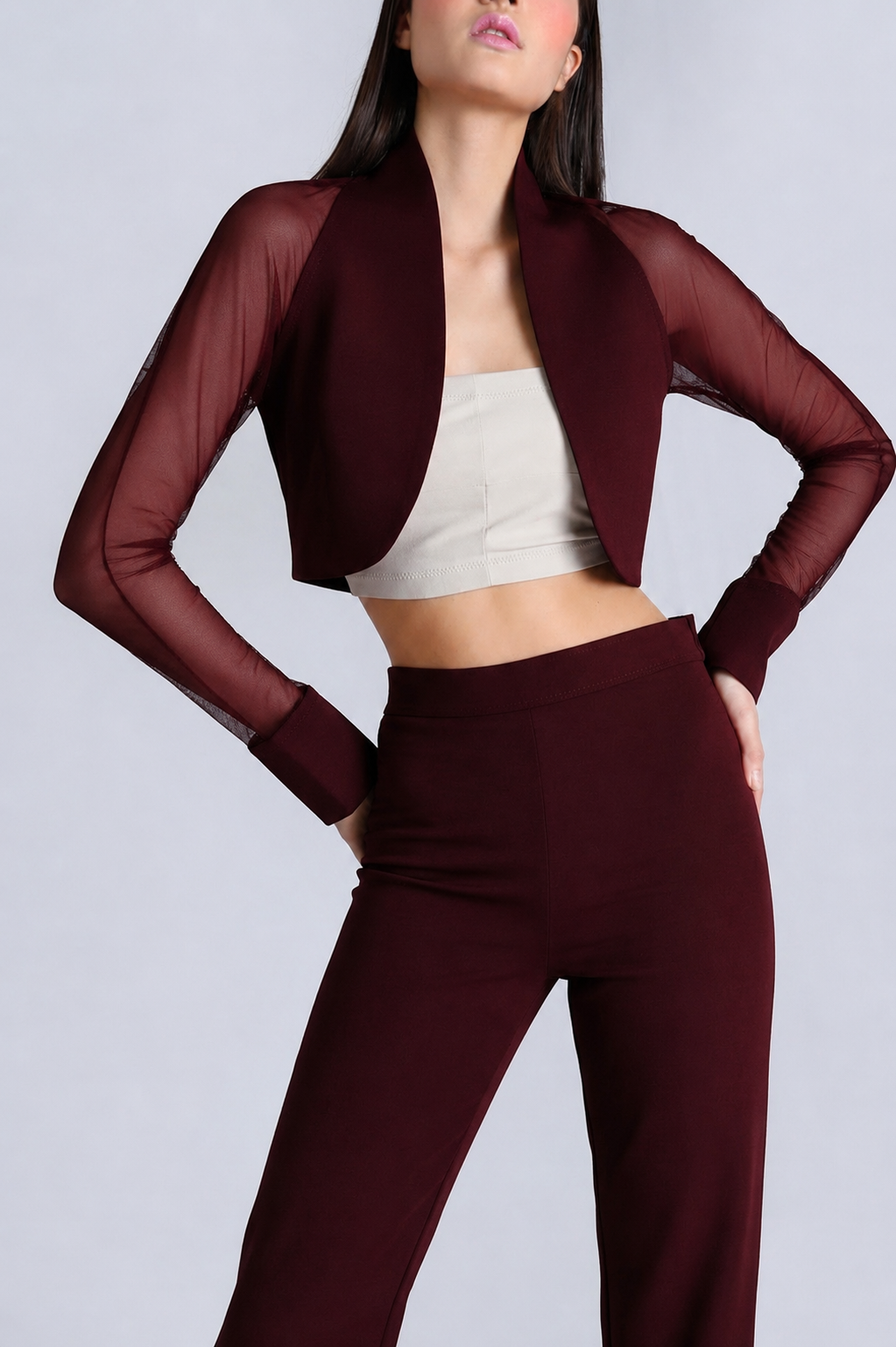A model wearing a burgundy two-piece set consisting of a cropped open-front jacket with sheer long sleeves and matching high-waisted trousers, styled over a beige crop top against a light gray studio background.