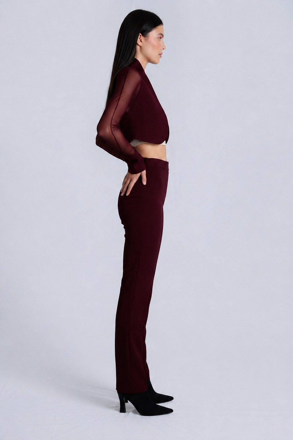 A woman stands in profile wearing a matching burgundy two-piece outfit consisting of a sheer long-sleeve crop top and high-waisted flared pants, paired with black pointed-toe heels, against a light gray studio background.