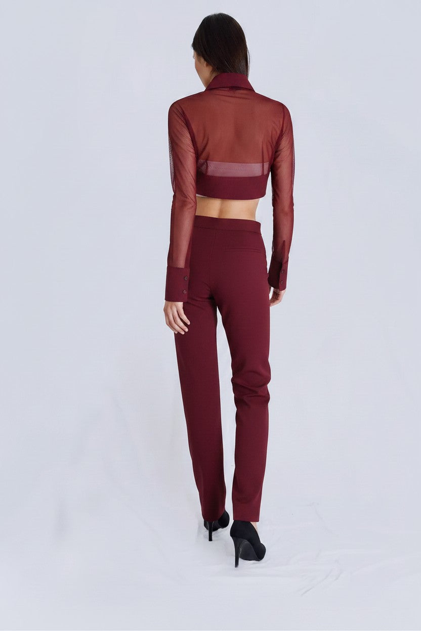 A model viewed from the back wearing a matching burgundy two-piece set consisting of a sheer long-sleeve crop top and high-waisted trousers, paired with black high heels on a light gray studio background.