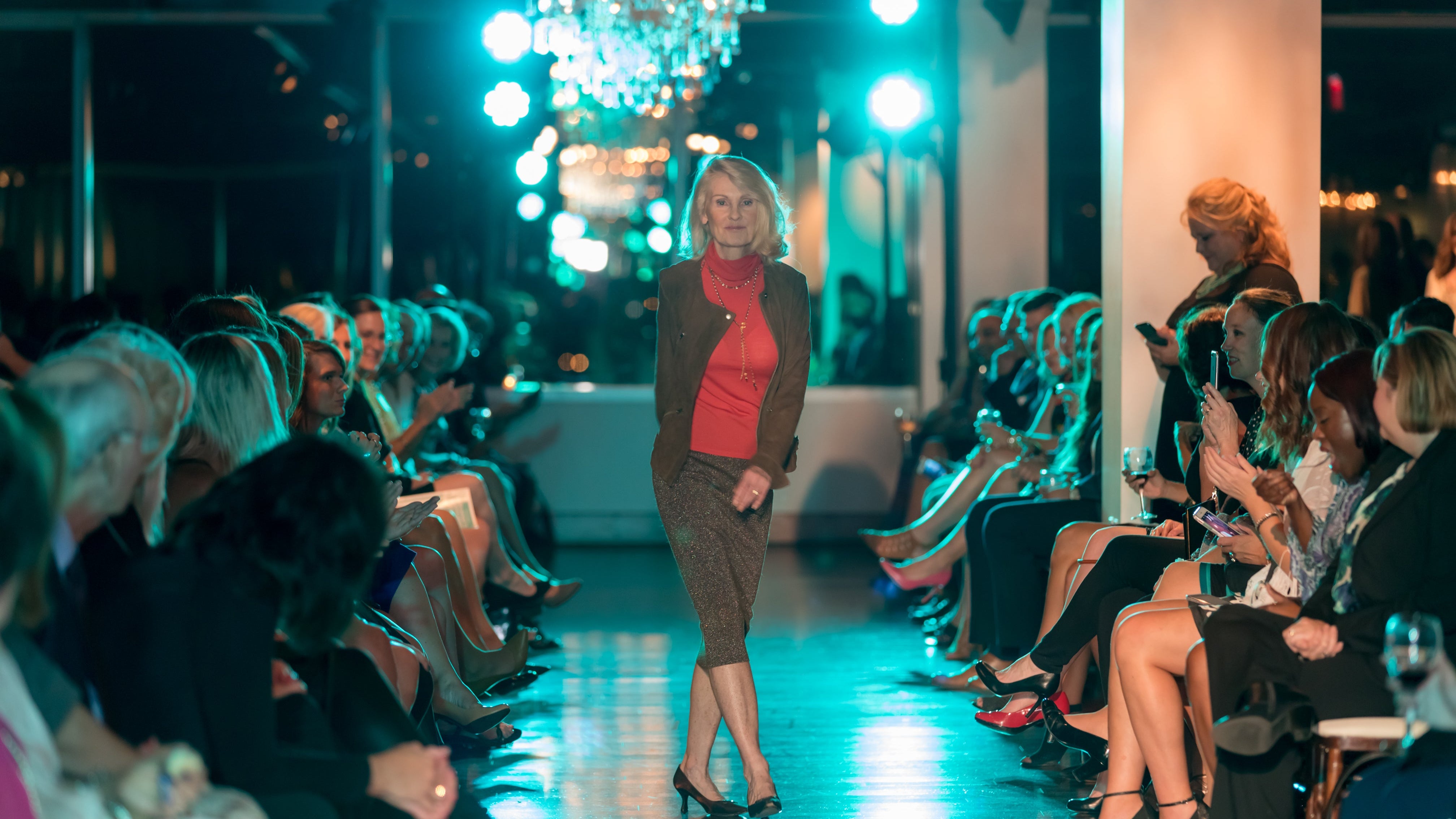 A woman walks down a fashion runway at a fashion show, wearing a dark brown blazer, a red top, and a brown patterned skirt. The runway is lined with an audience under bright stage lights with a chandelier hanging in the background.