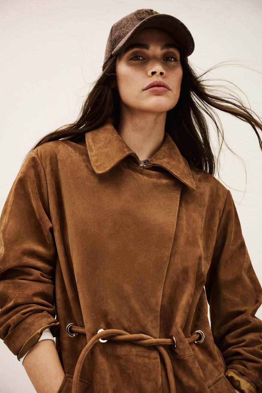 Close-up of a woman wearing a brown suede coat and cap against a neutral background.