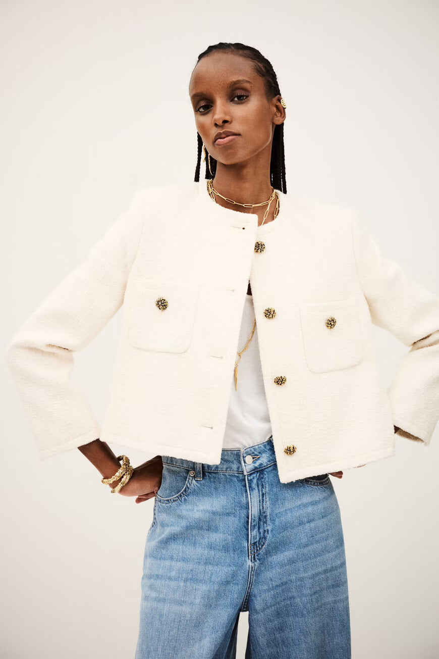 Woman wearing a white jacket with gold buttons and blue jeans on a plain background.