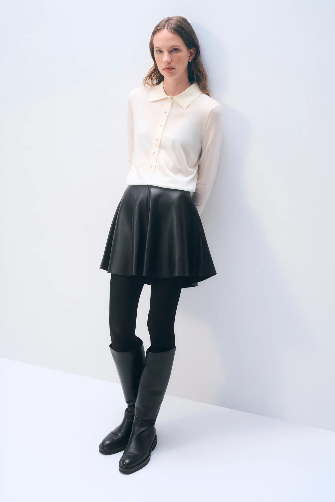 Woman wearing a cream blouse, black leather skirt, and black knee-high boots against a white background.