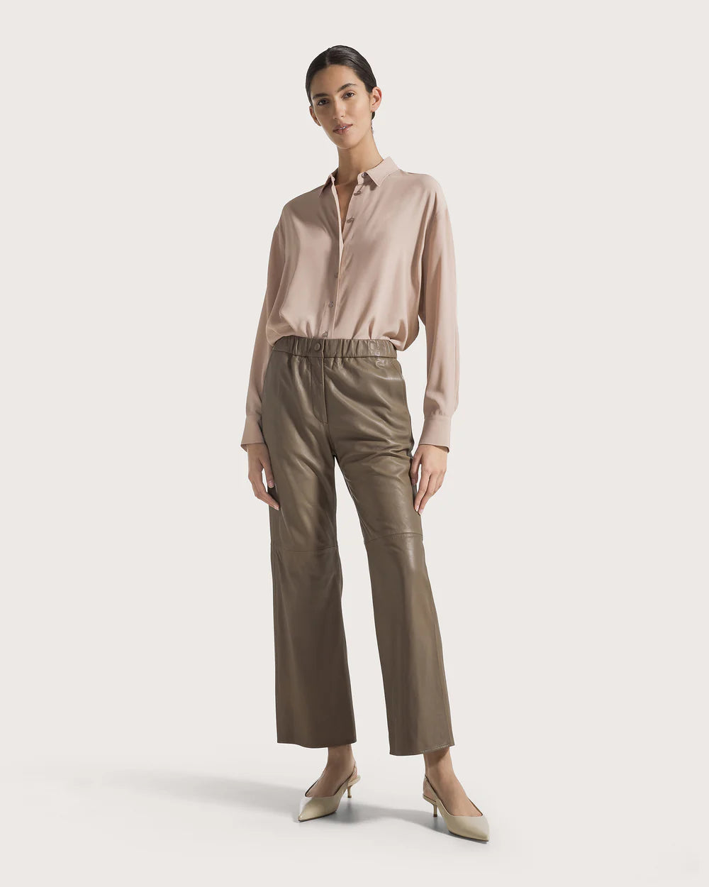 Woman wearing a beige blouse and taupe colored leather pants on a light background.