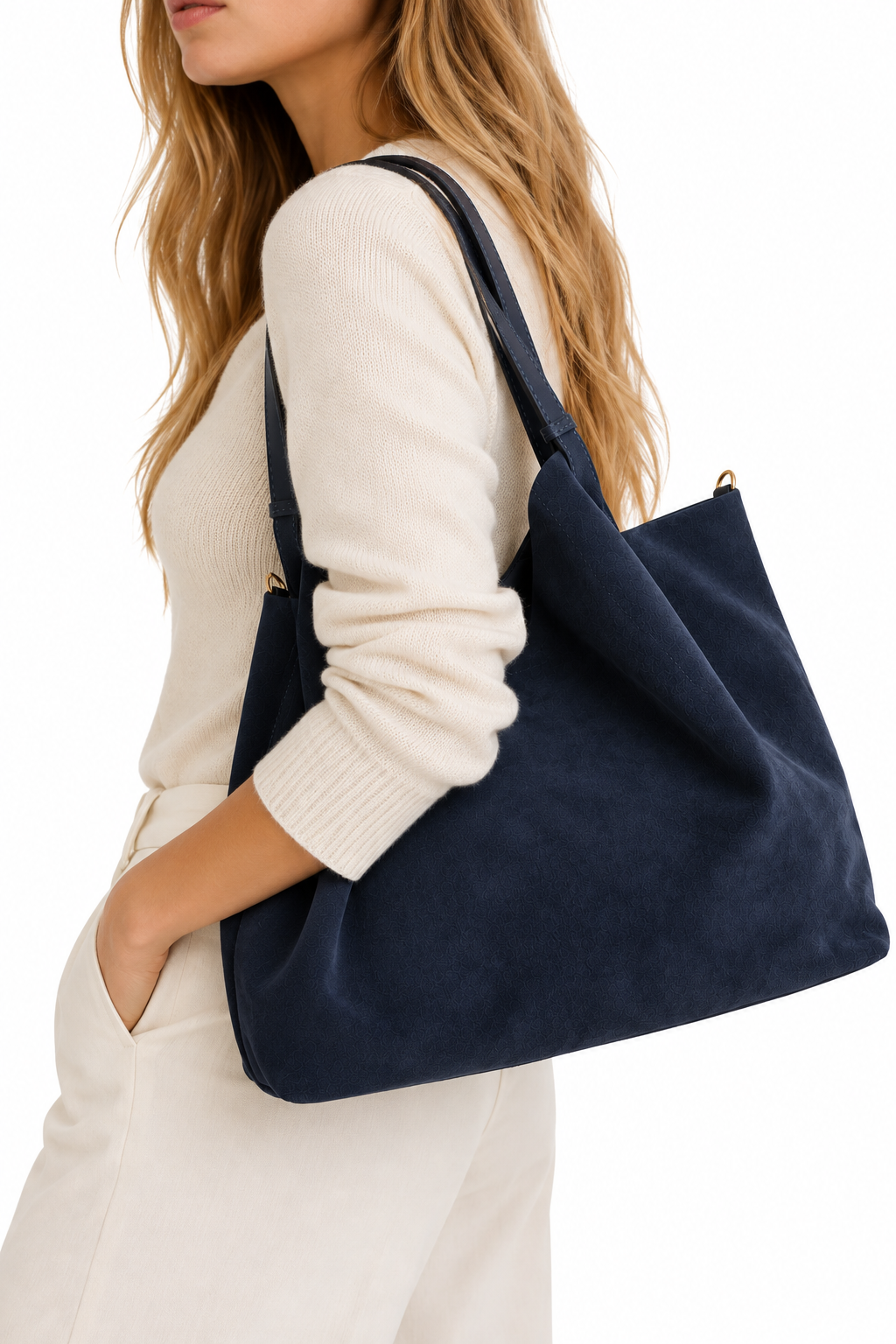 A woman wearing a cream ribbed sweater and cream pants carries a large navy blue suede tote bag over her shoulder, shown against a plain white background.