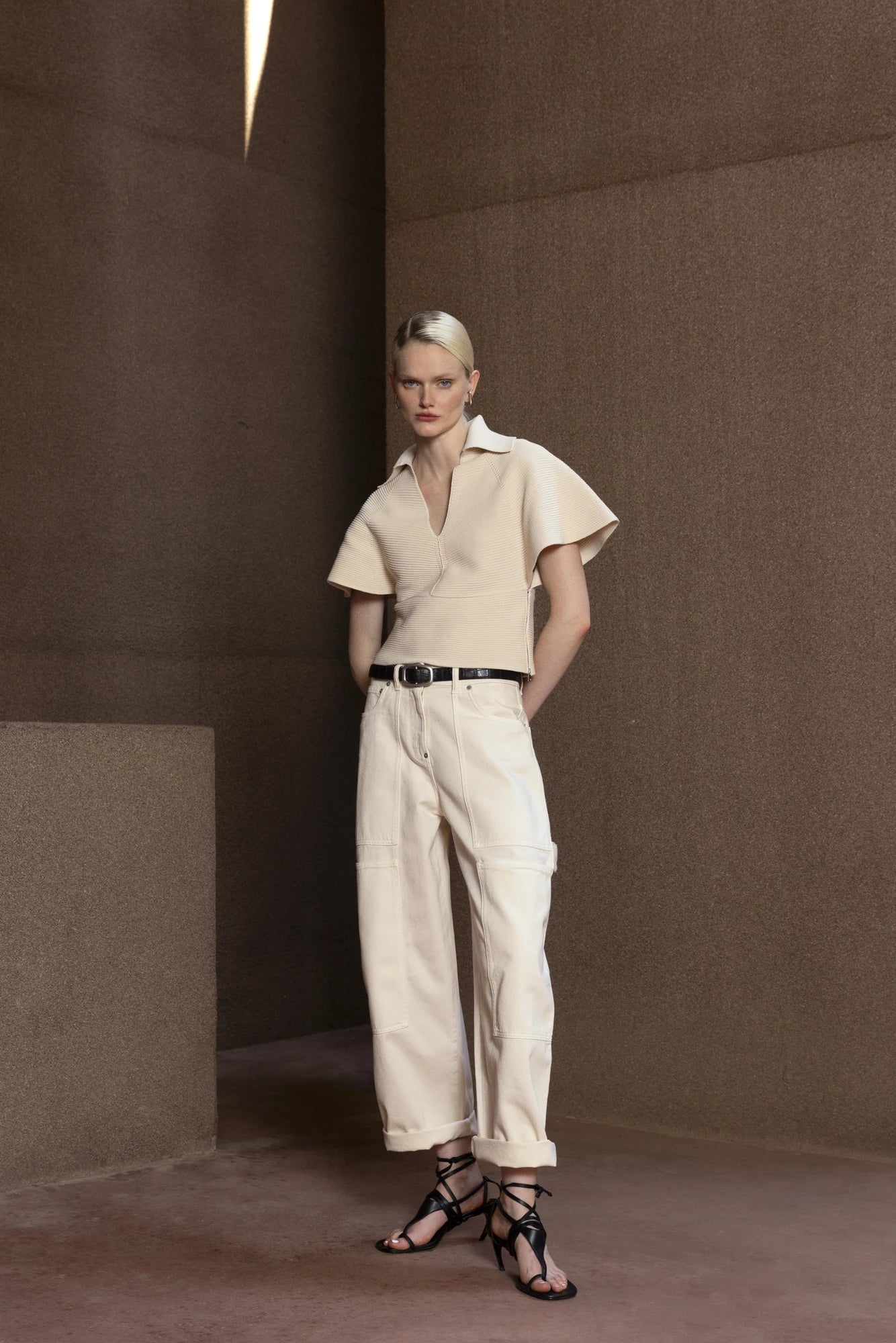 Woman in a beige shirt and white pants standing against a brown wall.