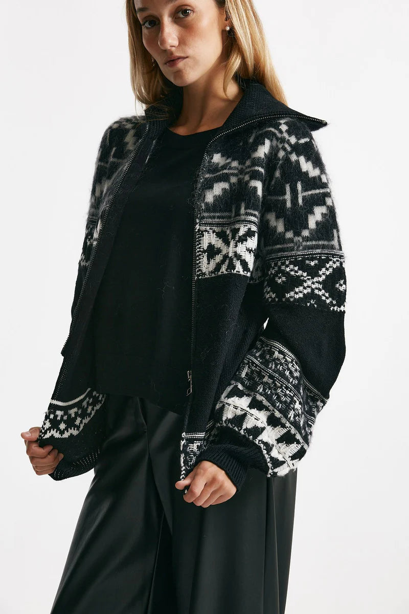 Woman wearing a black zip up sweater with white patterned sleeves on a white background.