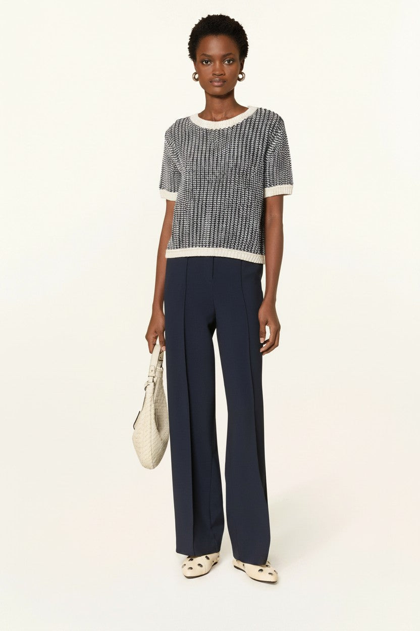 Woman wearing a off-white and bavy shortsleeve sweater with navy pants and cream shoes on a plain background.