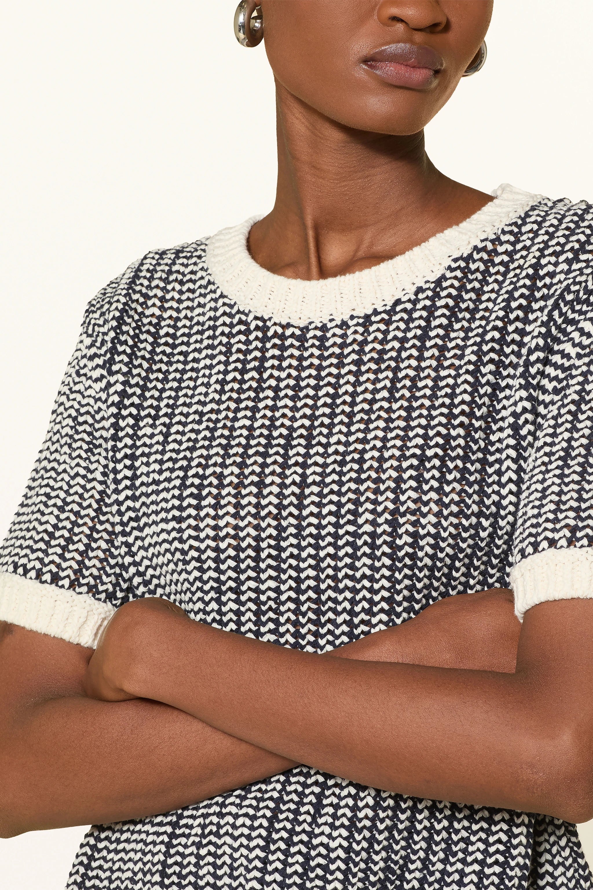 Close-up of a woman wearing a off-white and navy shortsleeve knit sweater on a plain background.