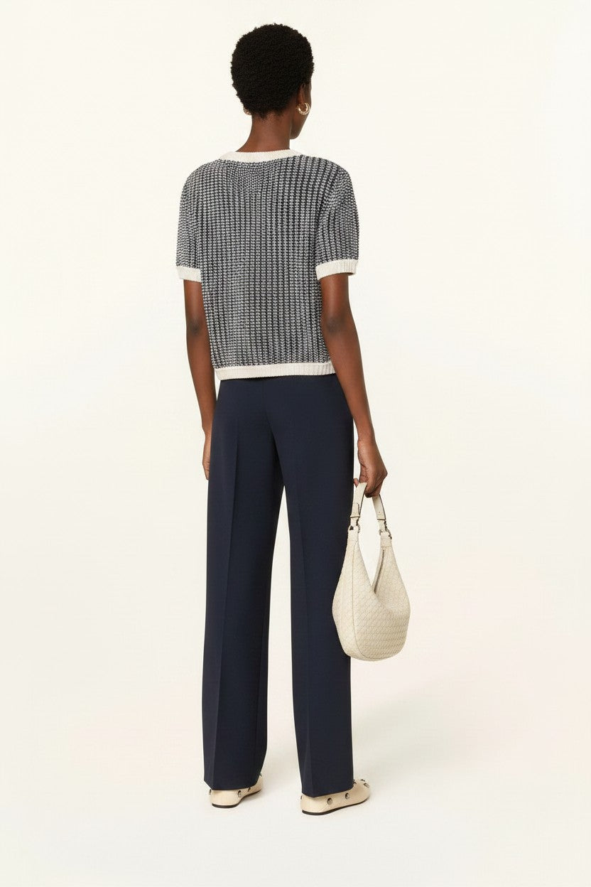 The back of a woman wearing a off-white and navy shortsleeve knit sweater with navy pants, cream shoes, and a cream bag on a plain background.